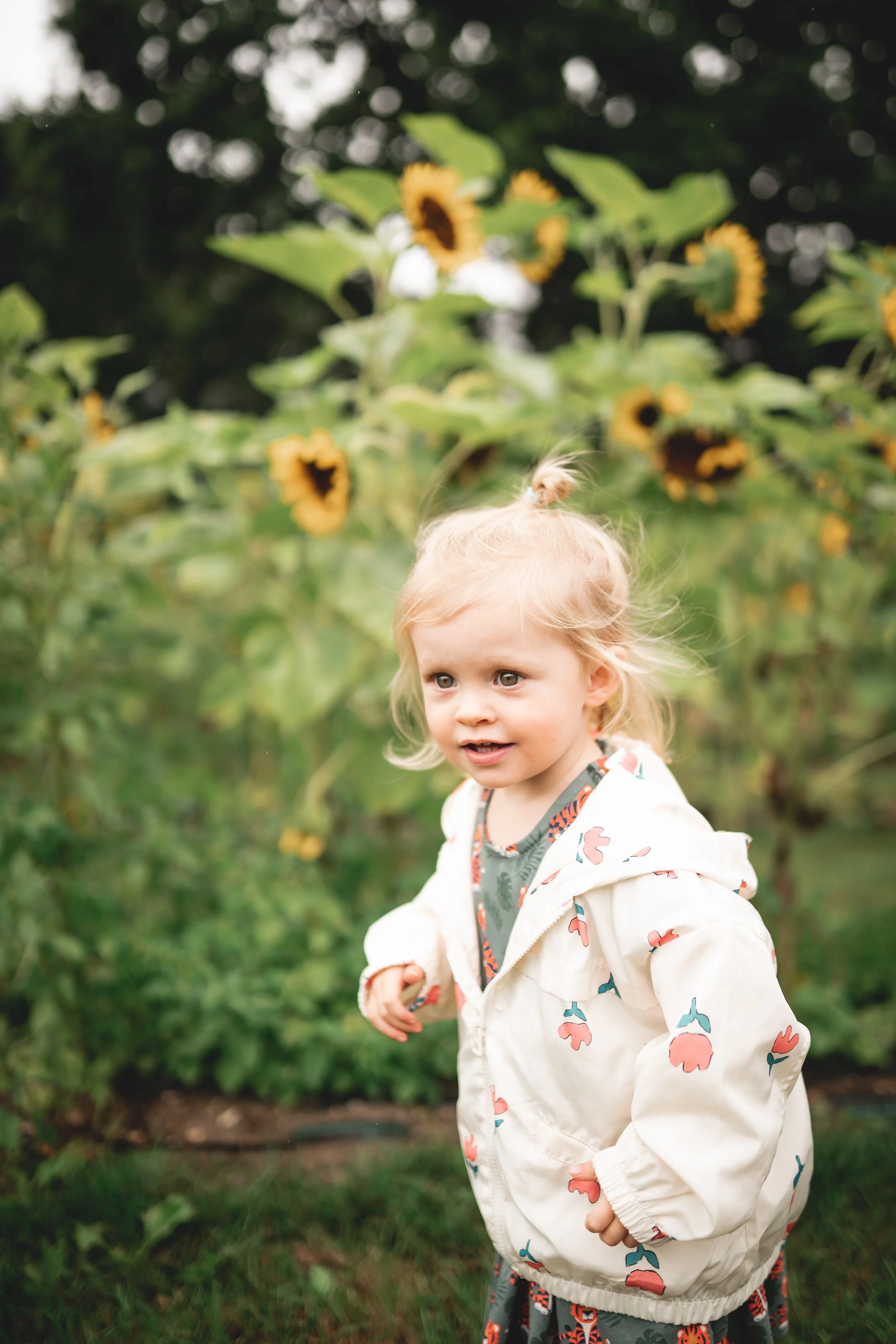 A young girl with blonde hair in a bun, wearing a white jacket with a floral pattern, stands outdoors in a garden with sunflowers in the background.