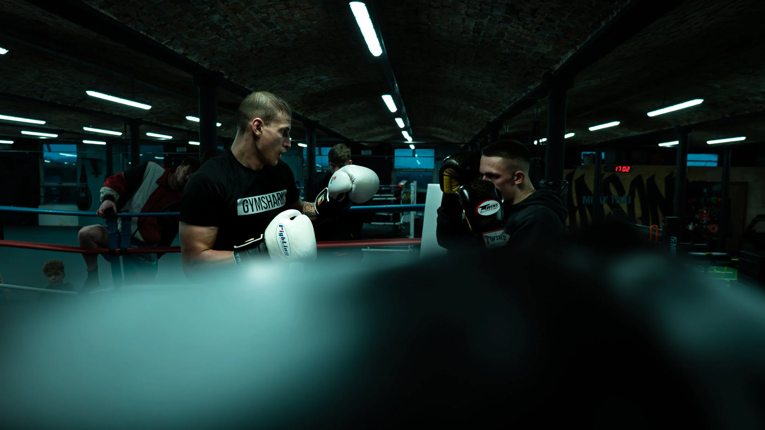 Two men in boxing gloves sparring inside a boxing ring with an overhead fluorescent light, with a person and a child watching in the background.