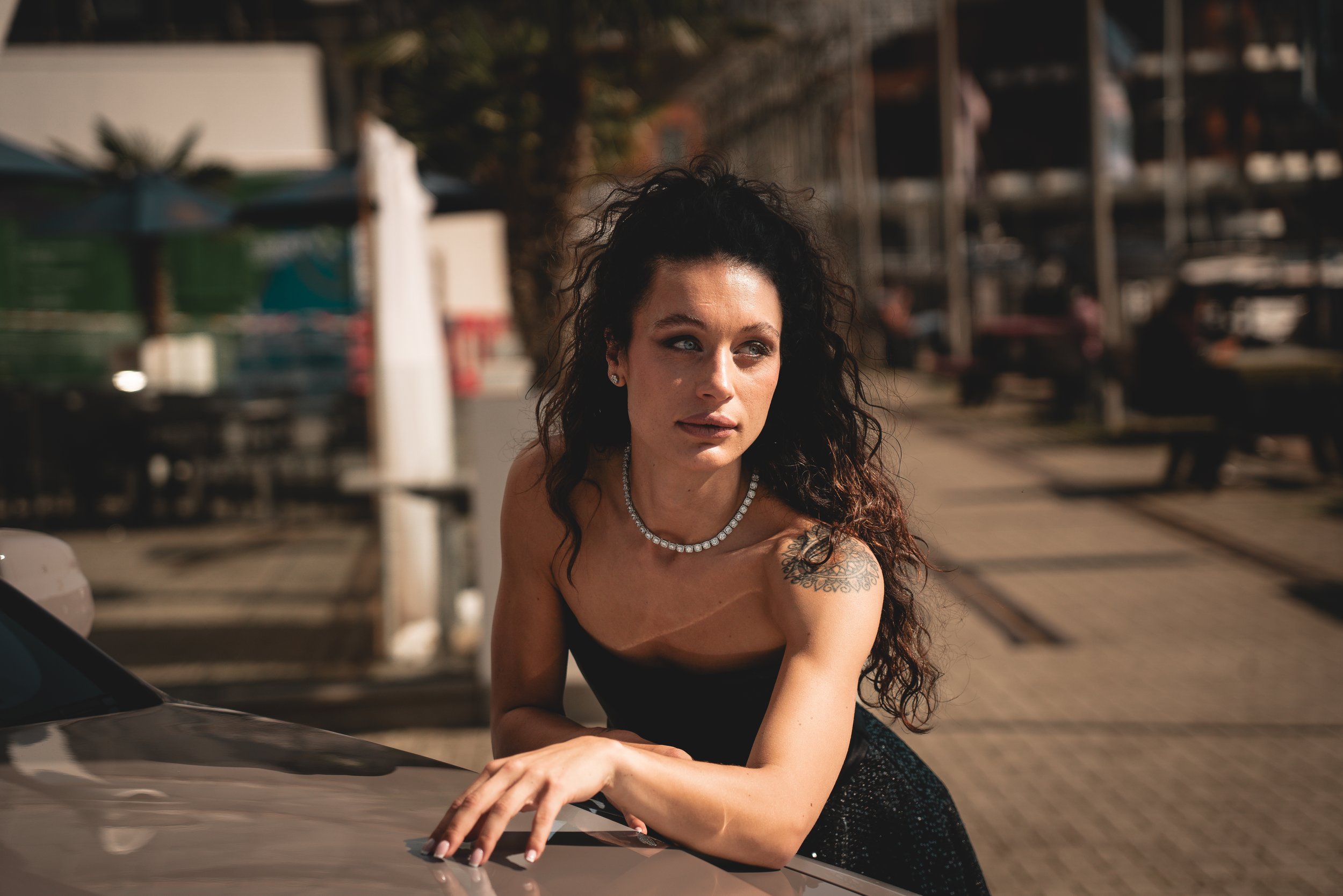 A woman with curly dark hair, wearing a black dress and a pearl necklace, leaning on a car in an outdoor setting during daytime.