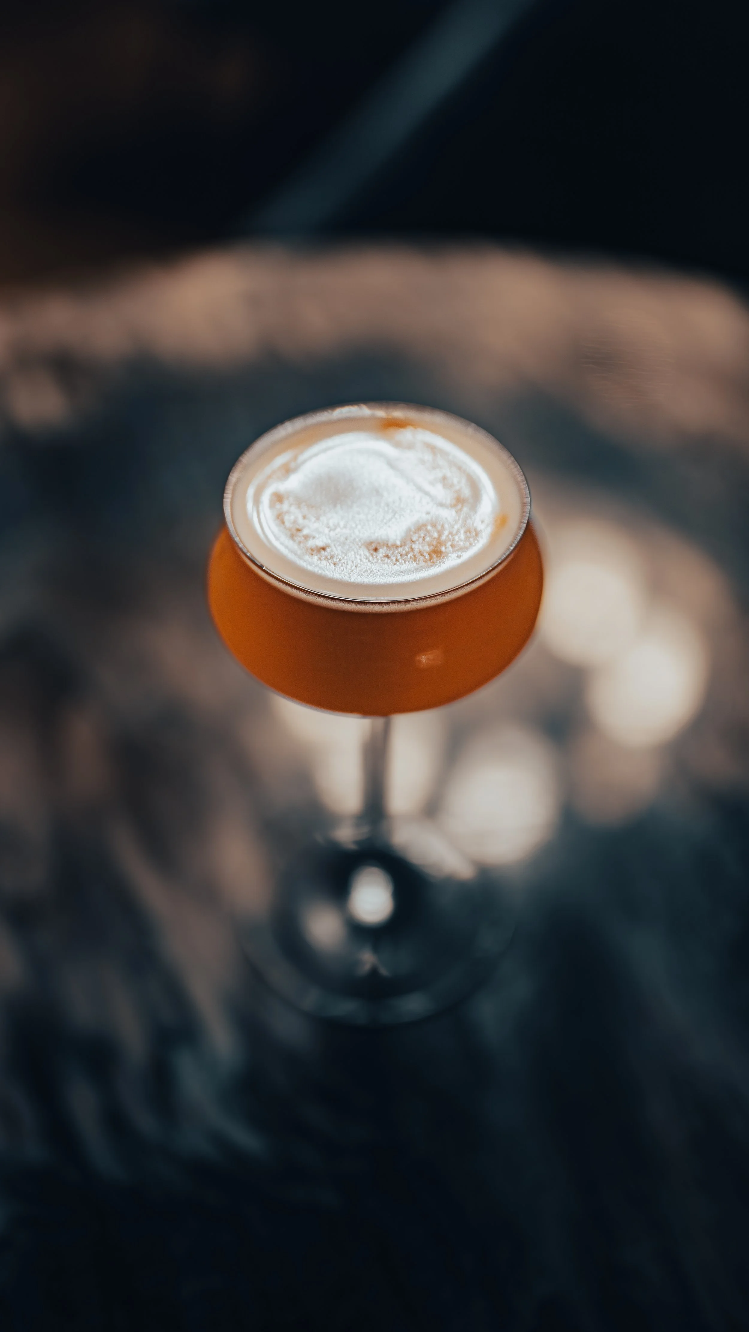A cocktail glass filled with an amber-colored drink topped with a dollop of white foam or whipped cream, set on a dark, textured surface.