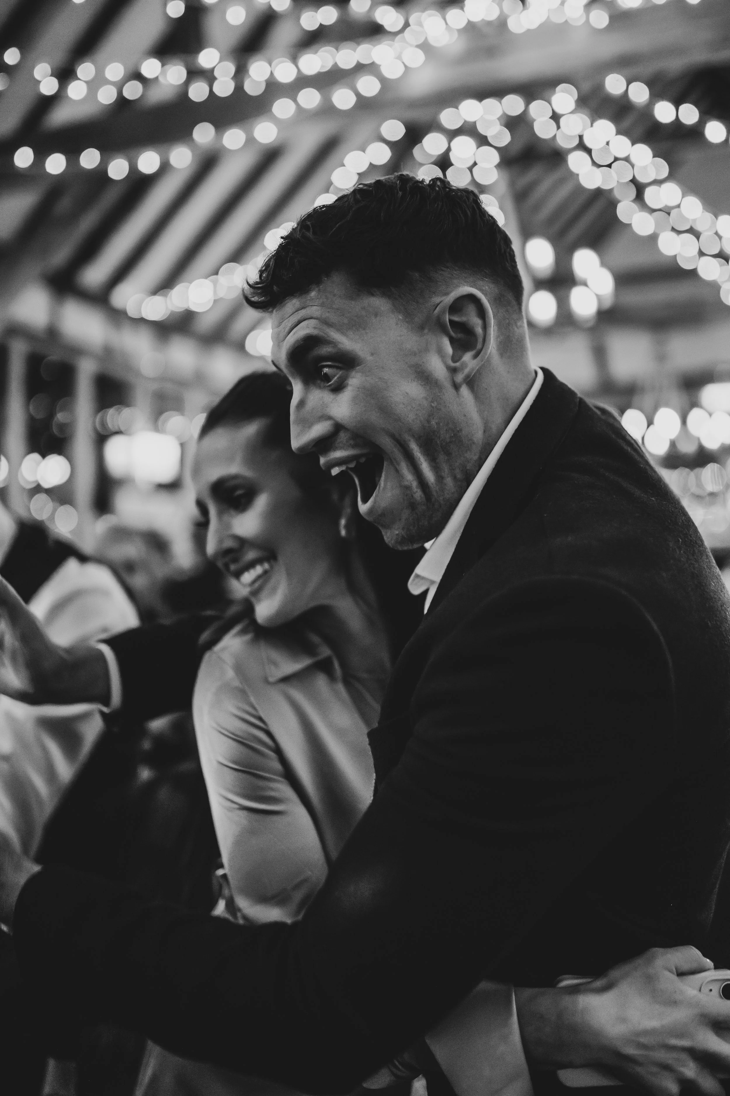 Two people, a man and a woman, smiling and enjoying a moment at a celebration with string lights overhead, black and white photo.