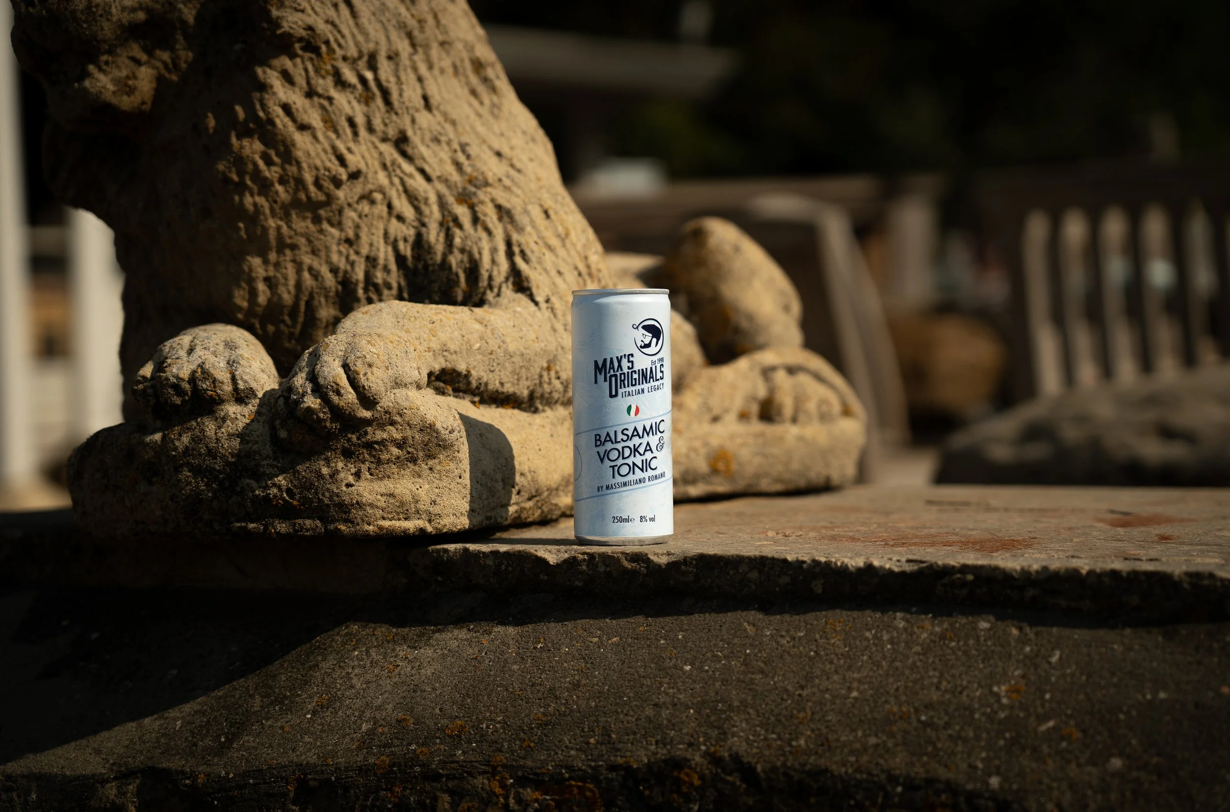 A can of Max's Originals Balsamic Vodka & Tonic drink placed on a weathered stone surface next to a stone sculpture of a lion's face and paws.