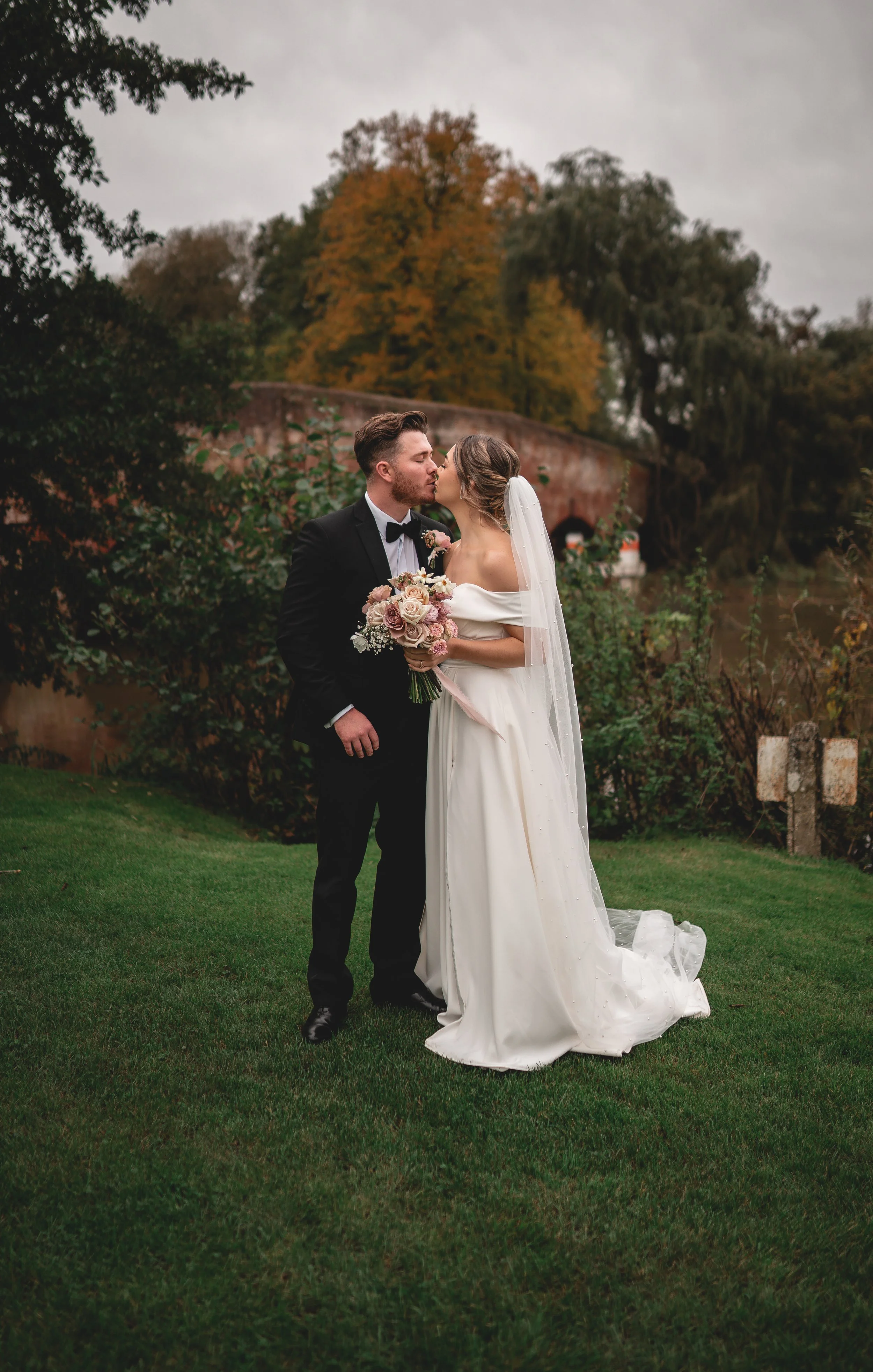 A newlywed couple sharing a kiss outdoors during their wedding, with the bride holding a bouquet of flowers and wearing a white gown and veil, and the groom in a black tuxedo, on a grassy lawn with autumn trees and a bridge in the background.