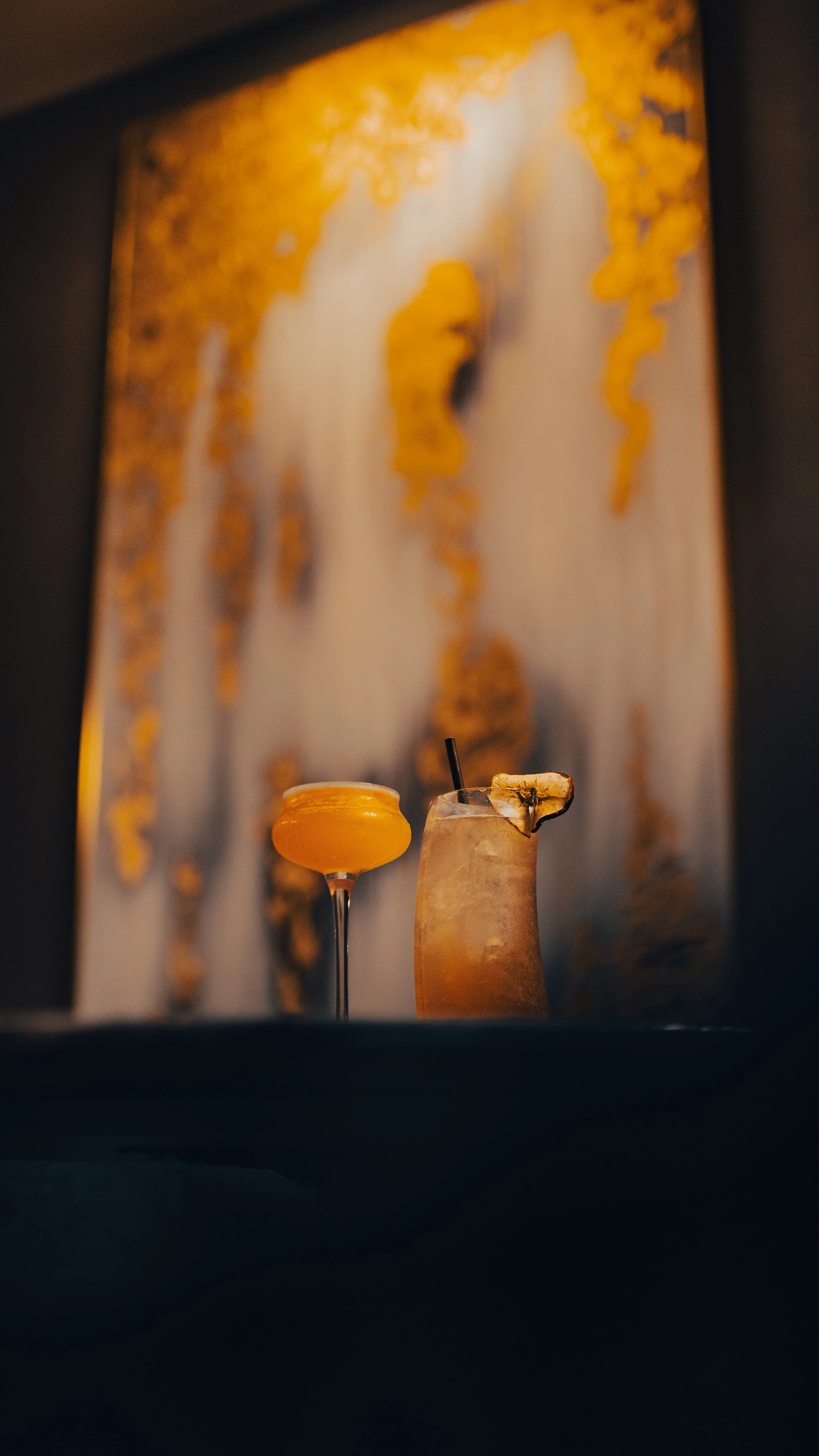 Two cocktails on a dark surface with a blurred abstract yellow and white artwork in the background. One cocktail is orange in a coupe glass, and the other is a dark-colored drink with a pineapple slice garnish and a straw.