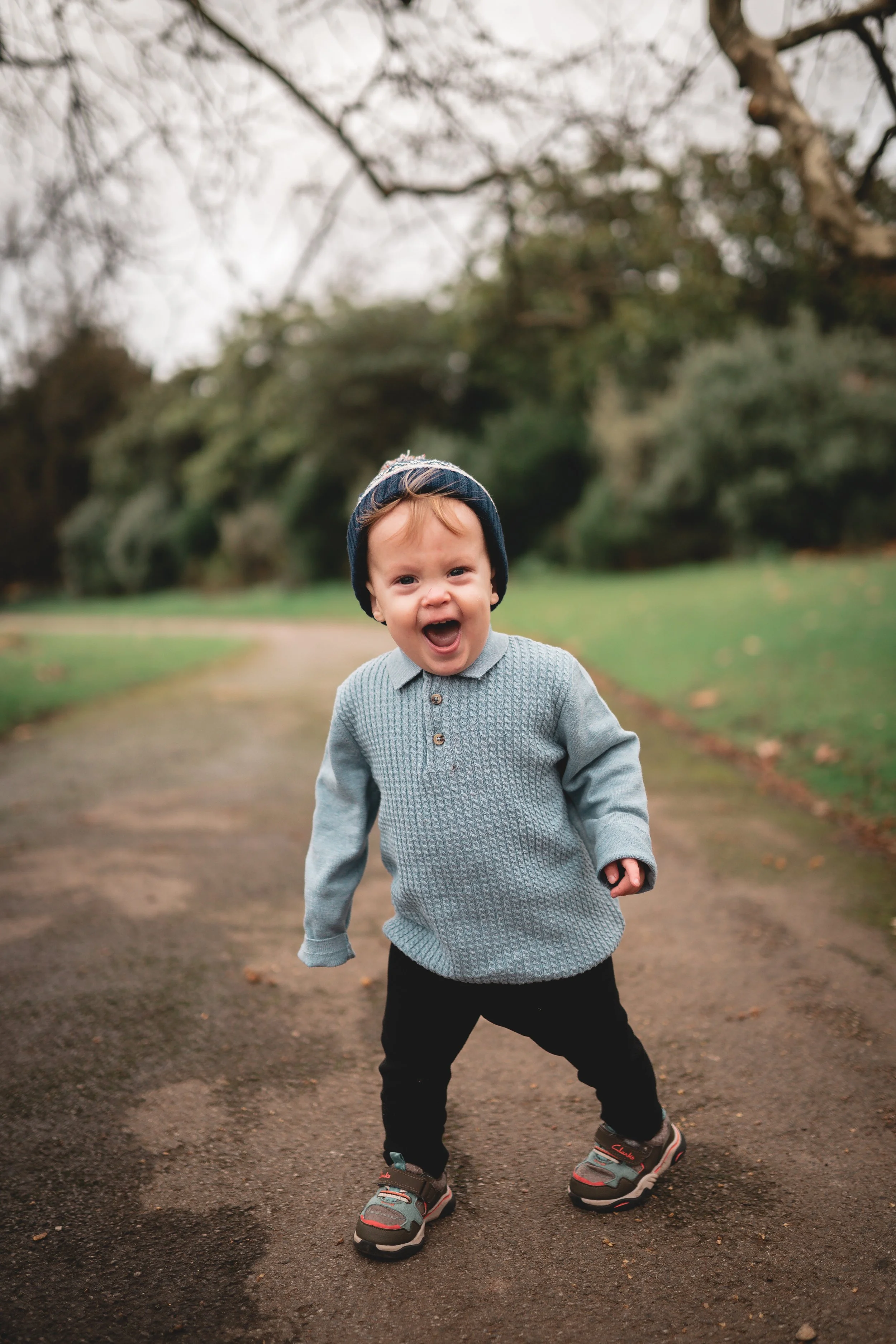 A young boy wearing a grey sweater, black pants, sneakers, and a beanie hat standing on a park path, smiling and shouting with arms slightly bent.