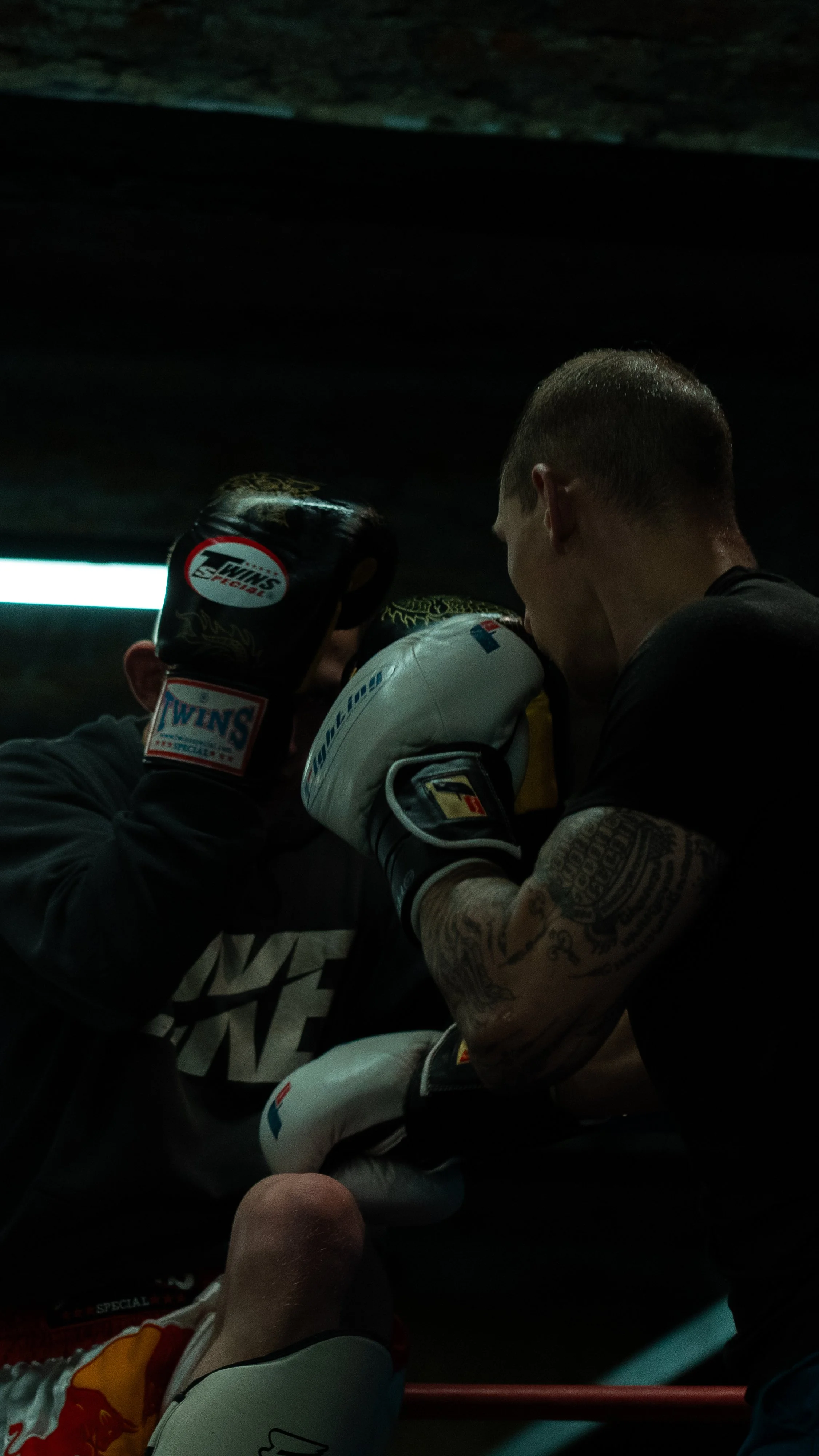 Two boxers in a training session, one wearing black gloves and the other wearing white gloves, practicing close combat under low-lit conditions.