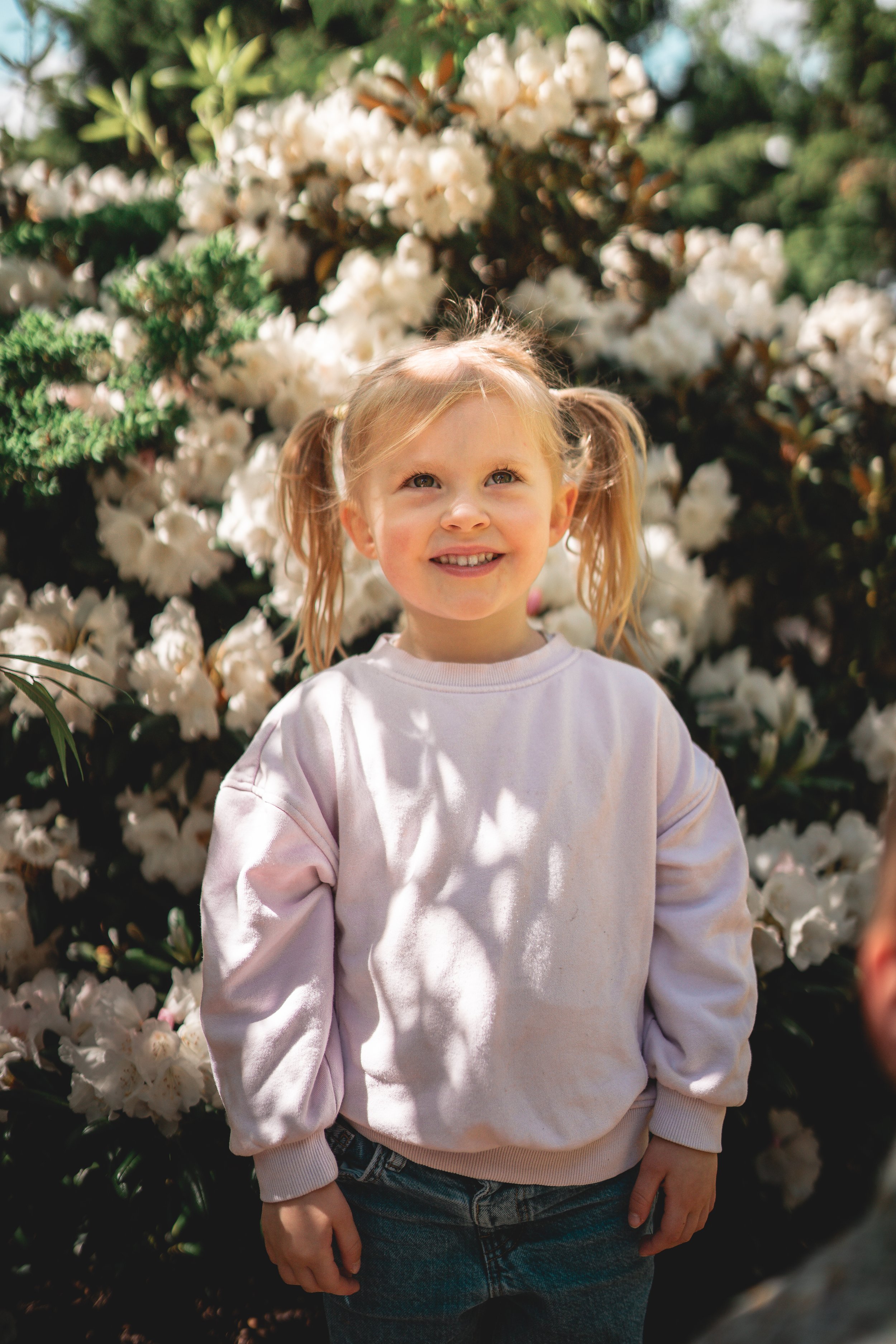 A young girl with blonde hair in pigtails, smiling and standing in front of blooming white flowers.