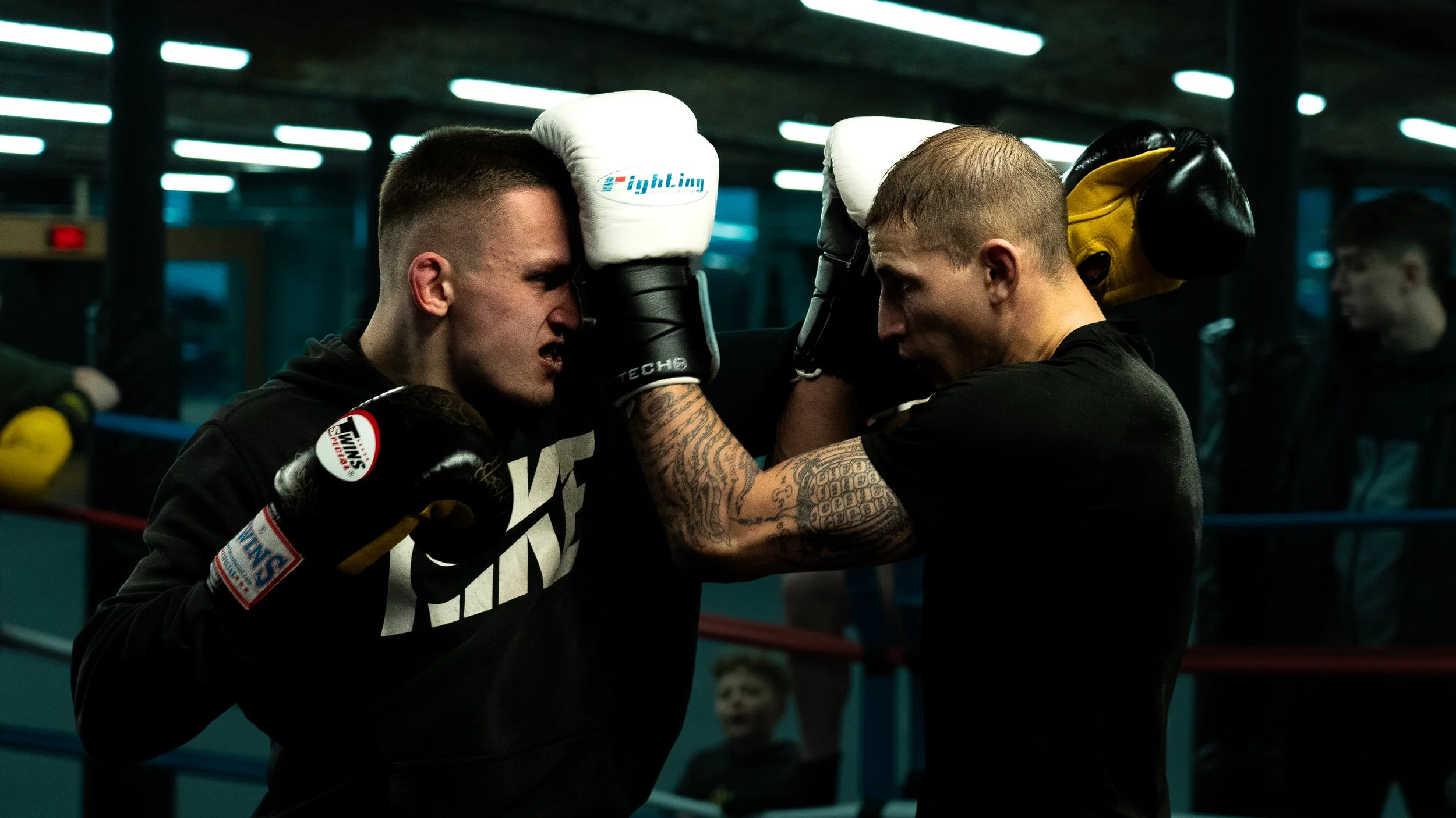 Two men in boxing gloves training in a boxing gym. One man is holding pads, and the other is in fighting stance, leaning forward cautiously.