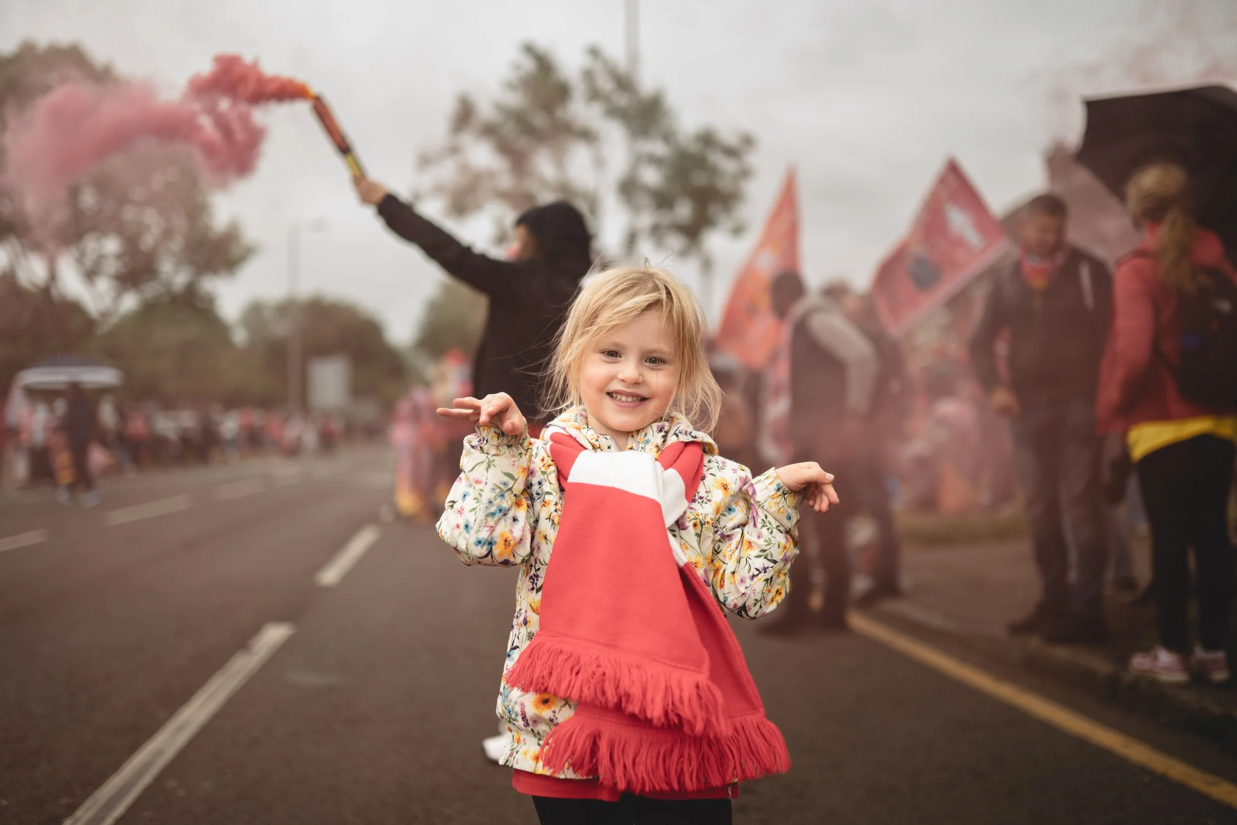 A young girl holding a red smoke flare during a parade or celebration, with people and flags in the background.