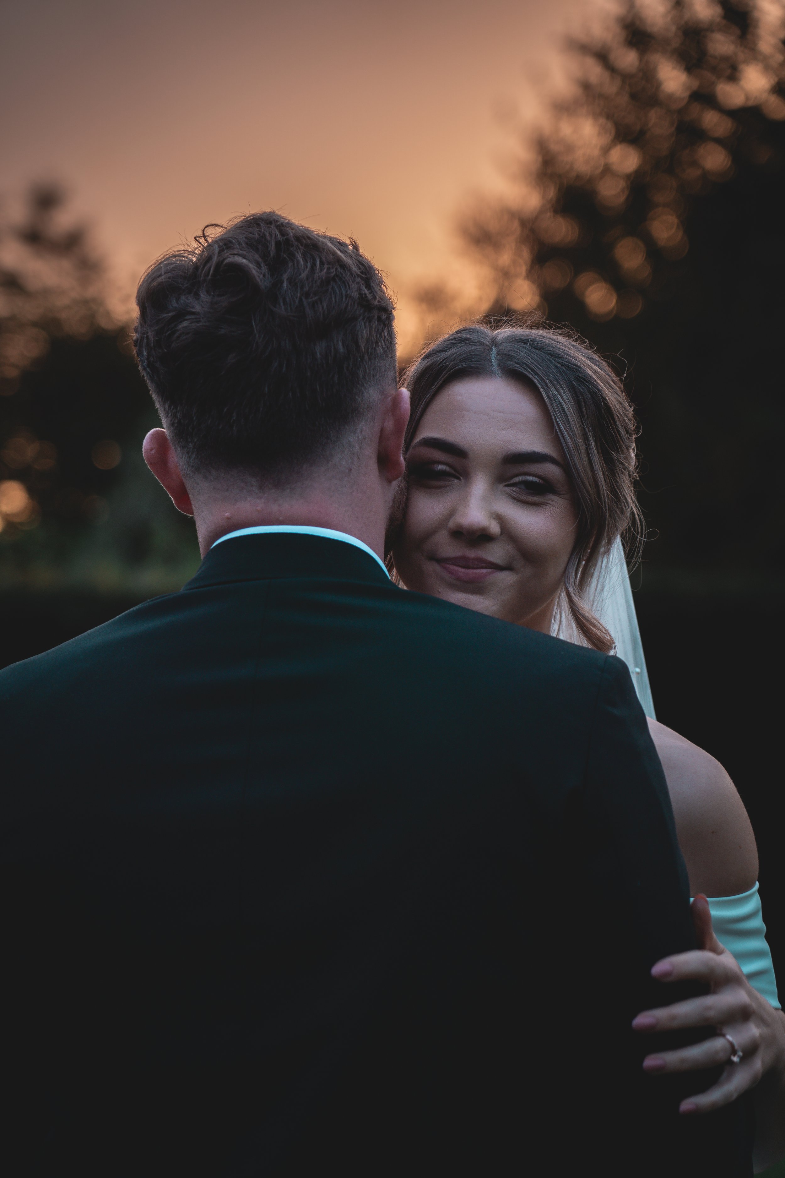 A bride and groom sharing a kiss outdoors at sunset, with the bride smiling and the groom facing away from the camera.