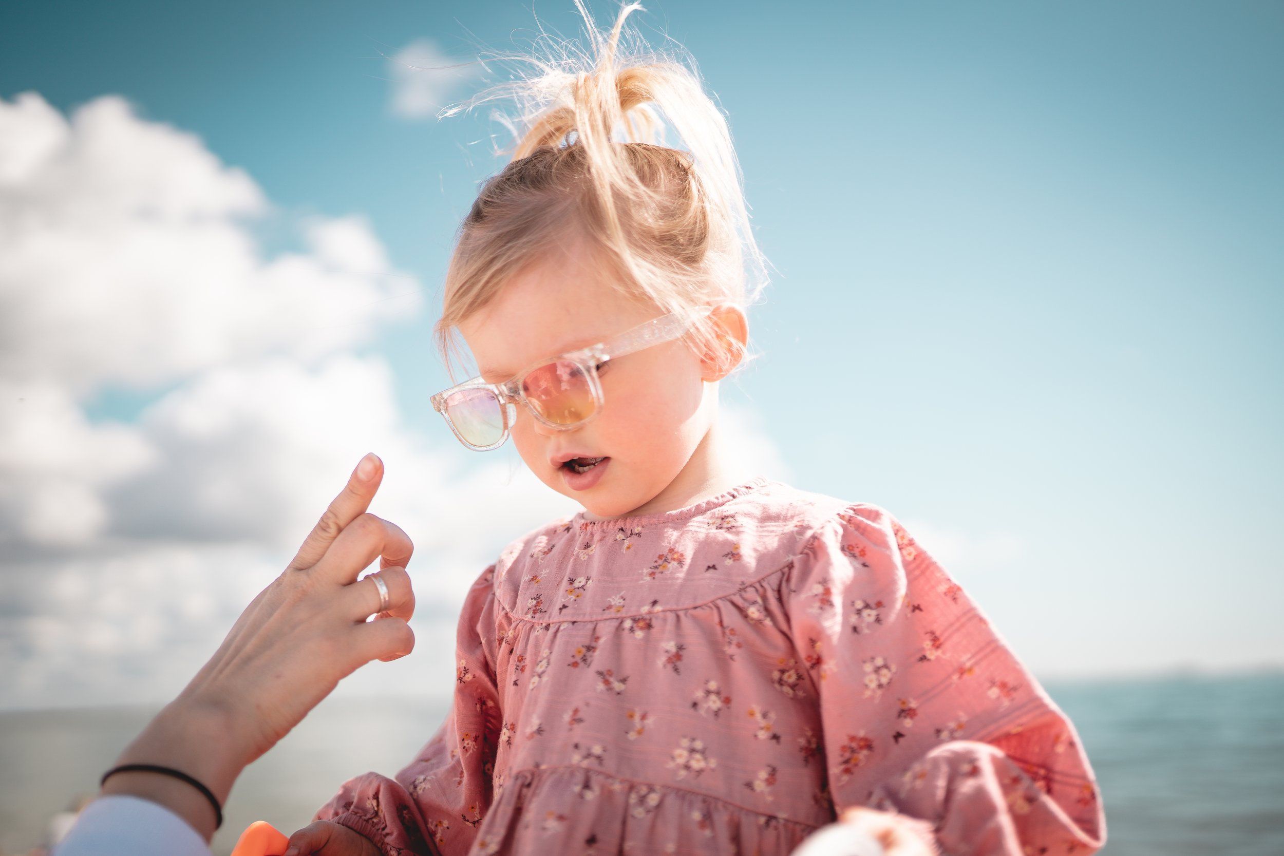 A young girl wearing pink floral dress and sunglasses on a sunny day outdoors near water, with a blue sky and clouds in the background.