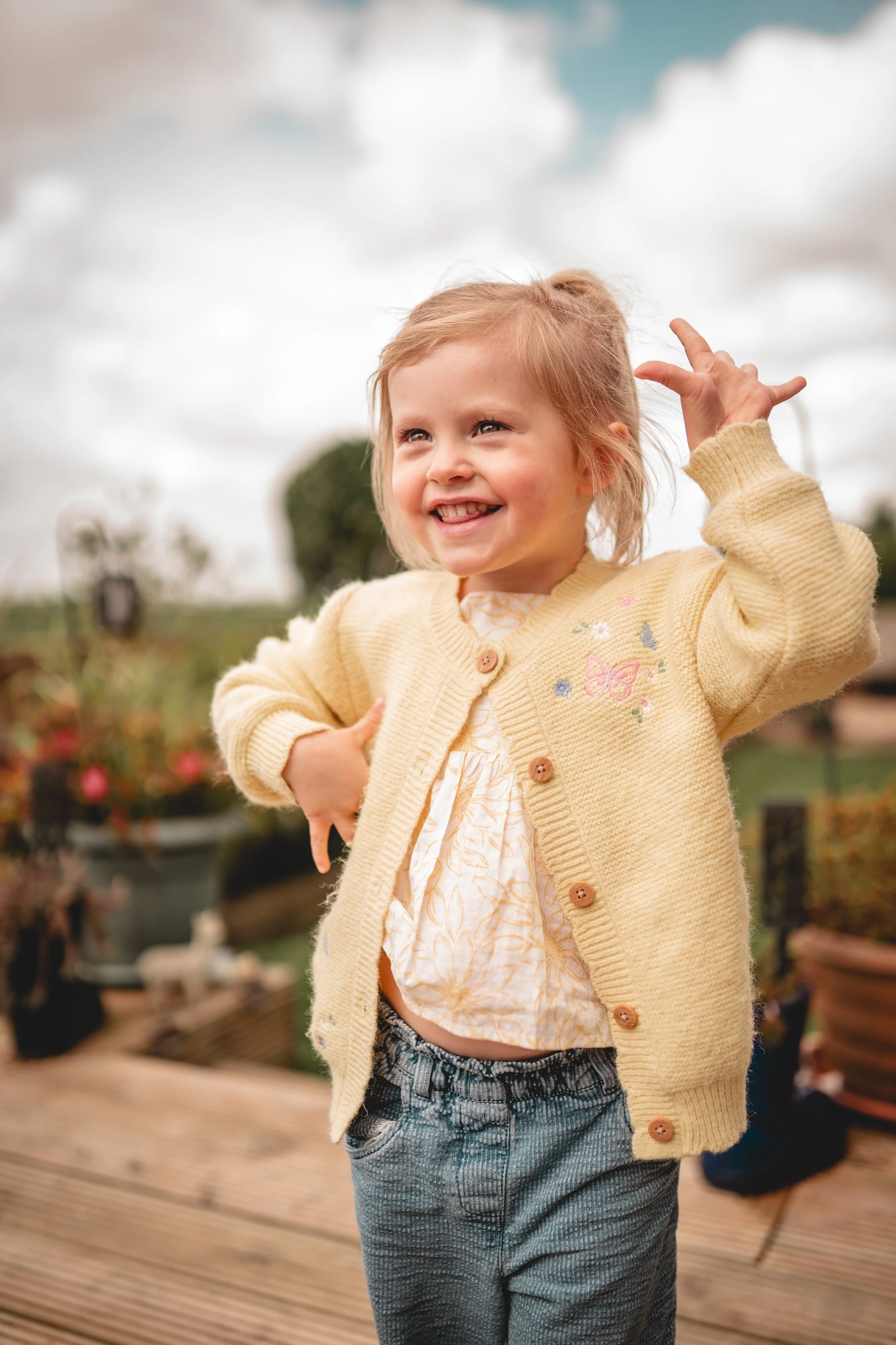 A young girl outside on a cloudy day, smiling and posing with her arms raised. She is wearing a yellow cardigan with embroidered flowers and butterfly, a patterned shirt, and gray pants.