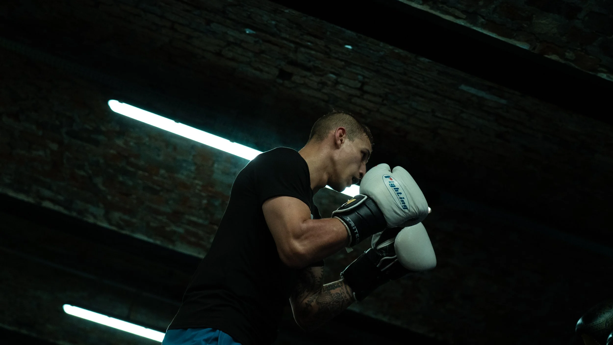 A male boxer wearing boxing gloves training in a gym with brick walls and fluorescent lighting.