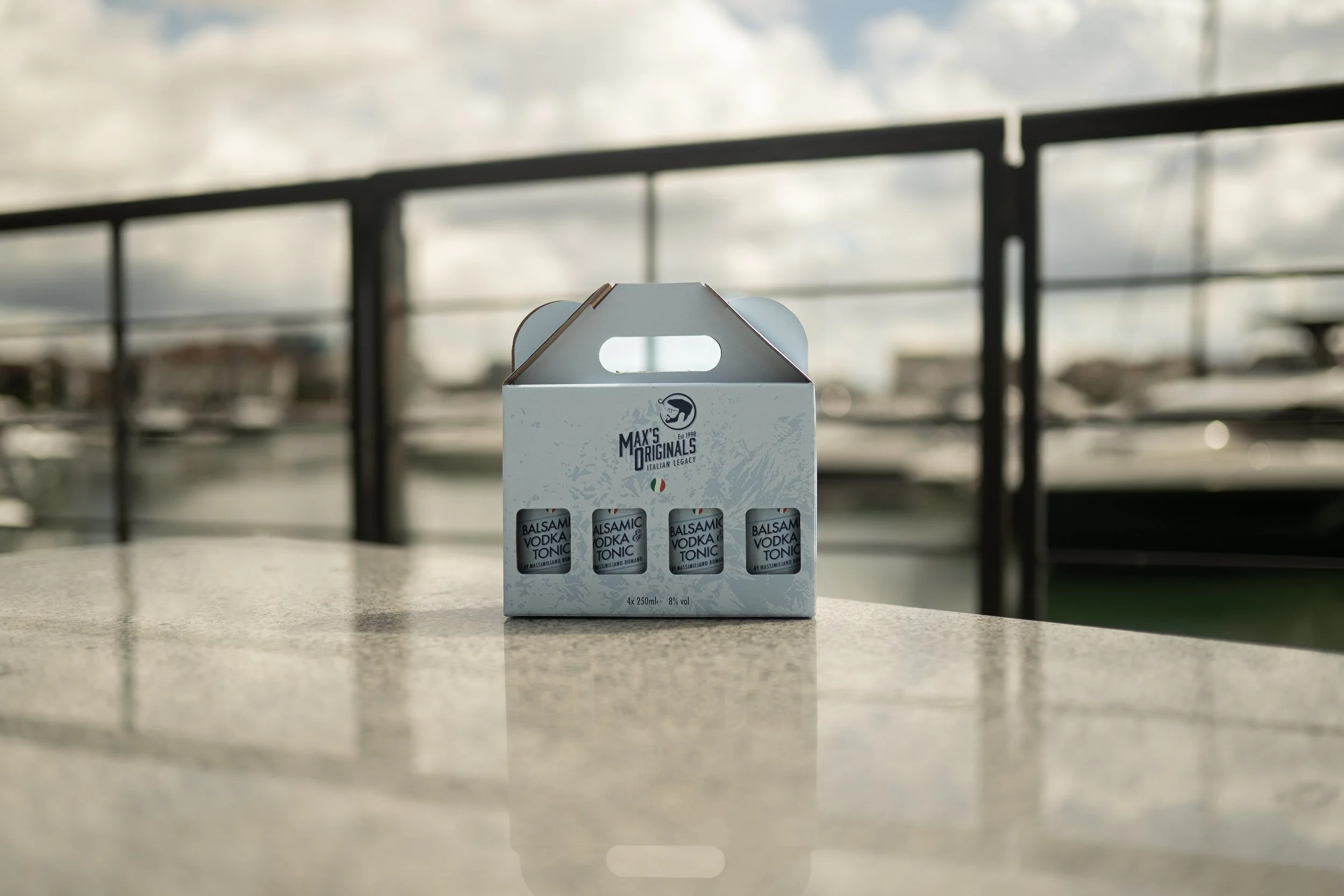 A white box labeled 'Max's Originals' with a small graphic and the words 'Italian Legacy' on it, containing four small bottles labeled 'Balsamic Vodka & Tonic' on a light-colored table with a blurry outdoor background.