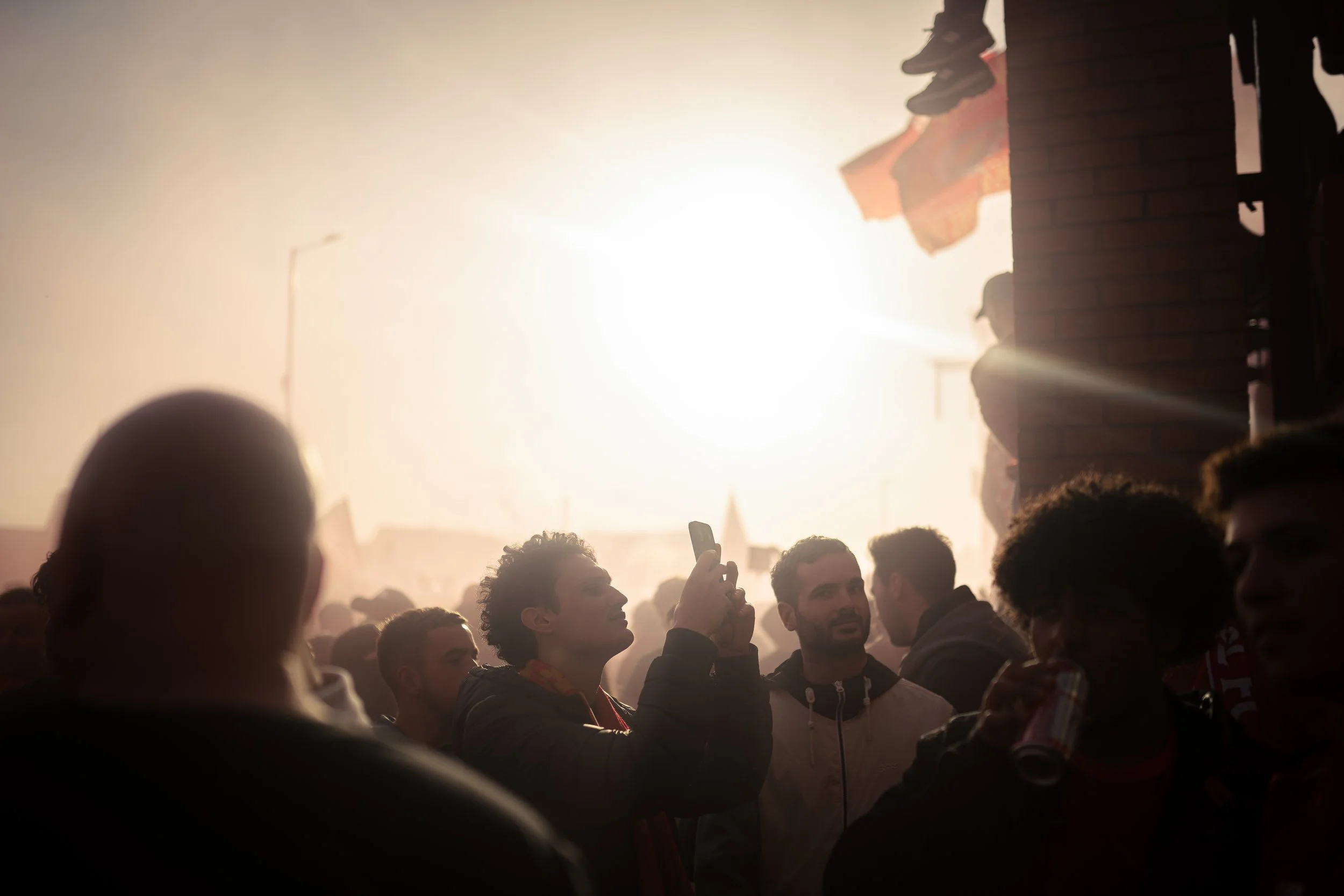 Group of young people at an outdoor gathering during sunset, some smiling and one person taking a photo with a smartphone, with clothes hanging on a line and a brick wall on the right.