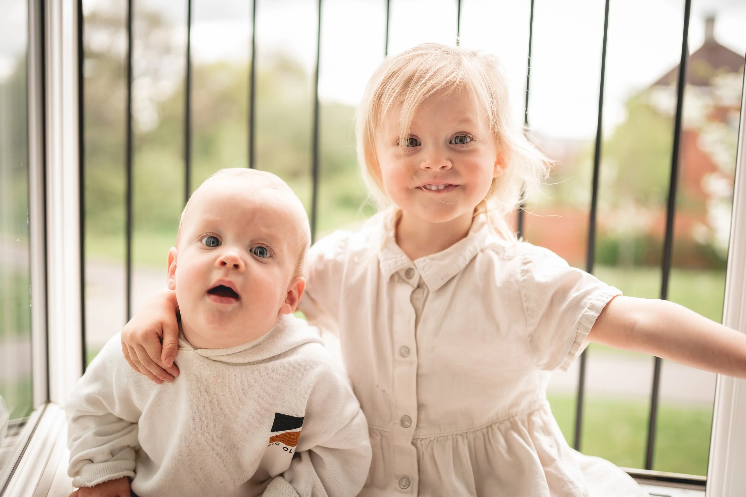 Two young children, a blonde girl and a baby boy, sitting on a windowsill with a view of a suburban neighborhood in the background. The girl has her arm around the boy, both smiling at the camera.