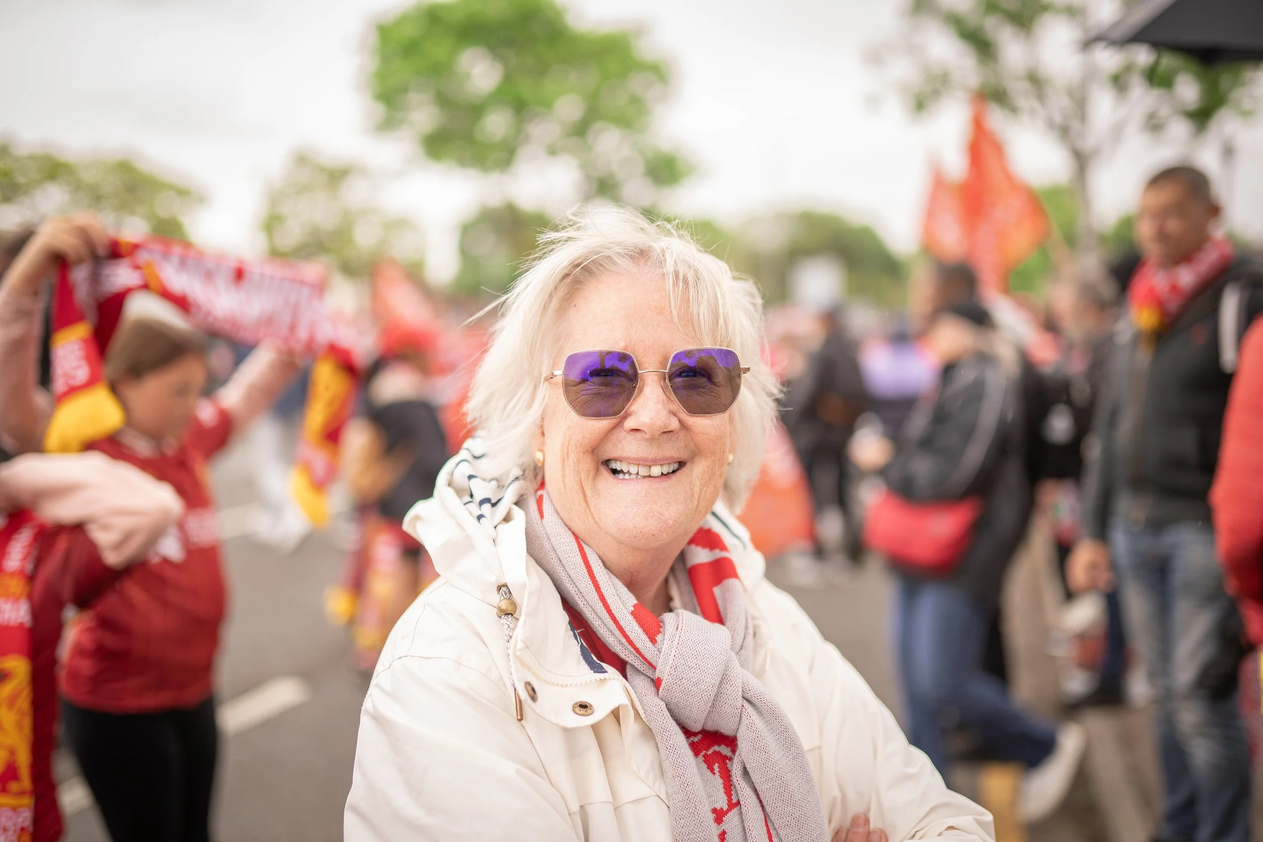 A cheerful elderly woman with blonde hair and purple sunglasses smiling at a public gathering or event, with people in the background holding red and yellow flags, some wearing red clothing, outdoors on a cloudy day.