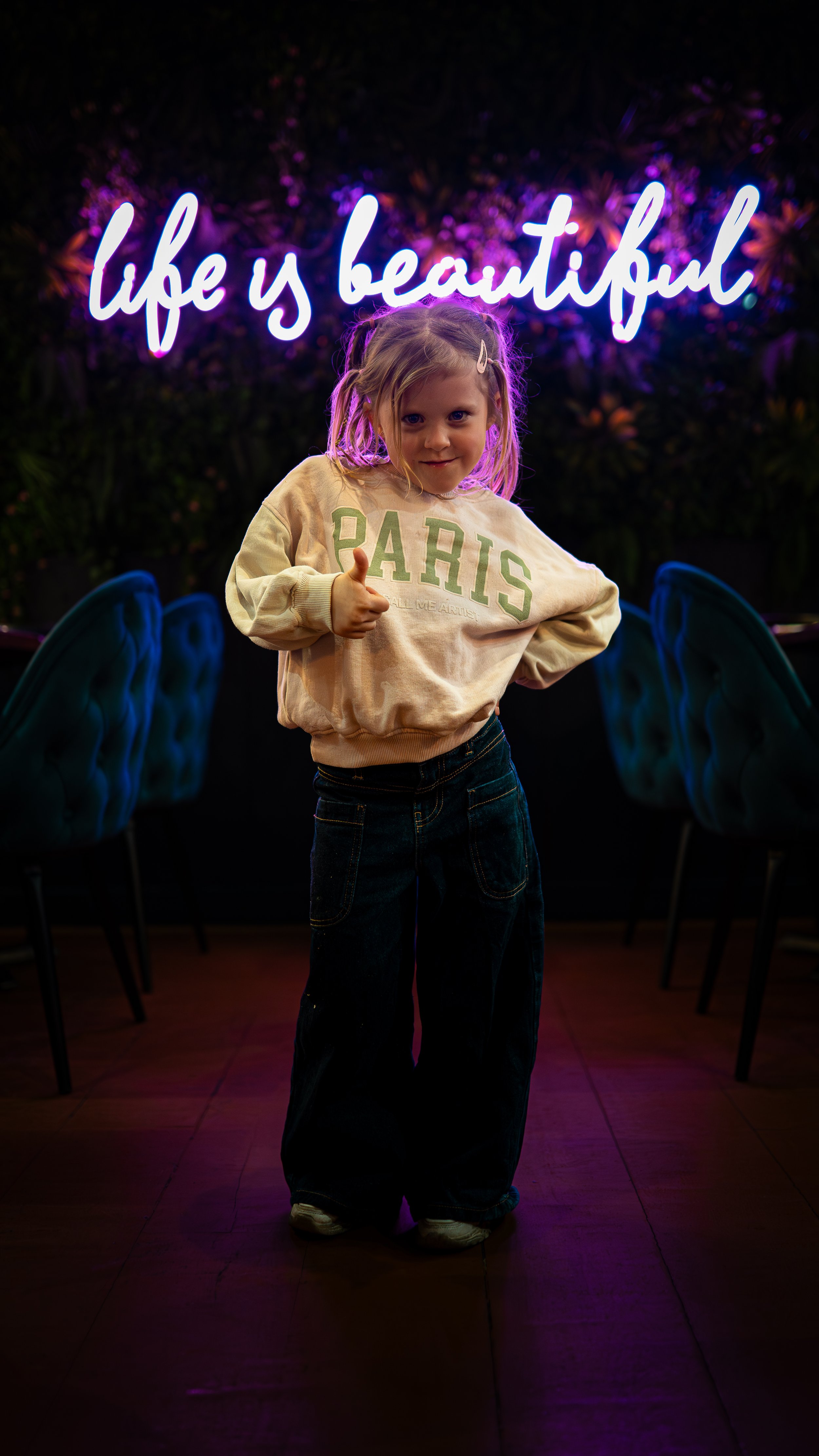 A young girl with blonde hair and a hair clip, wearing a cream sweatshirt with 'PARIS' written on it, giving a thumbs-up and smiling in front of a neon sign that says 'Life is beautiful' in cursive, with dark chairs in the background.