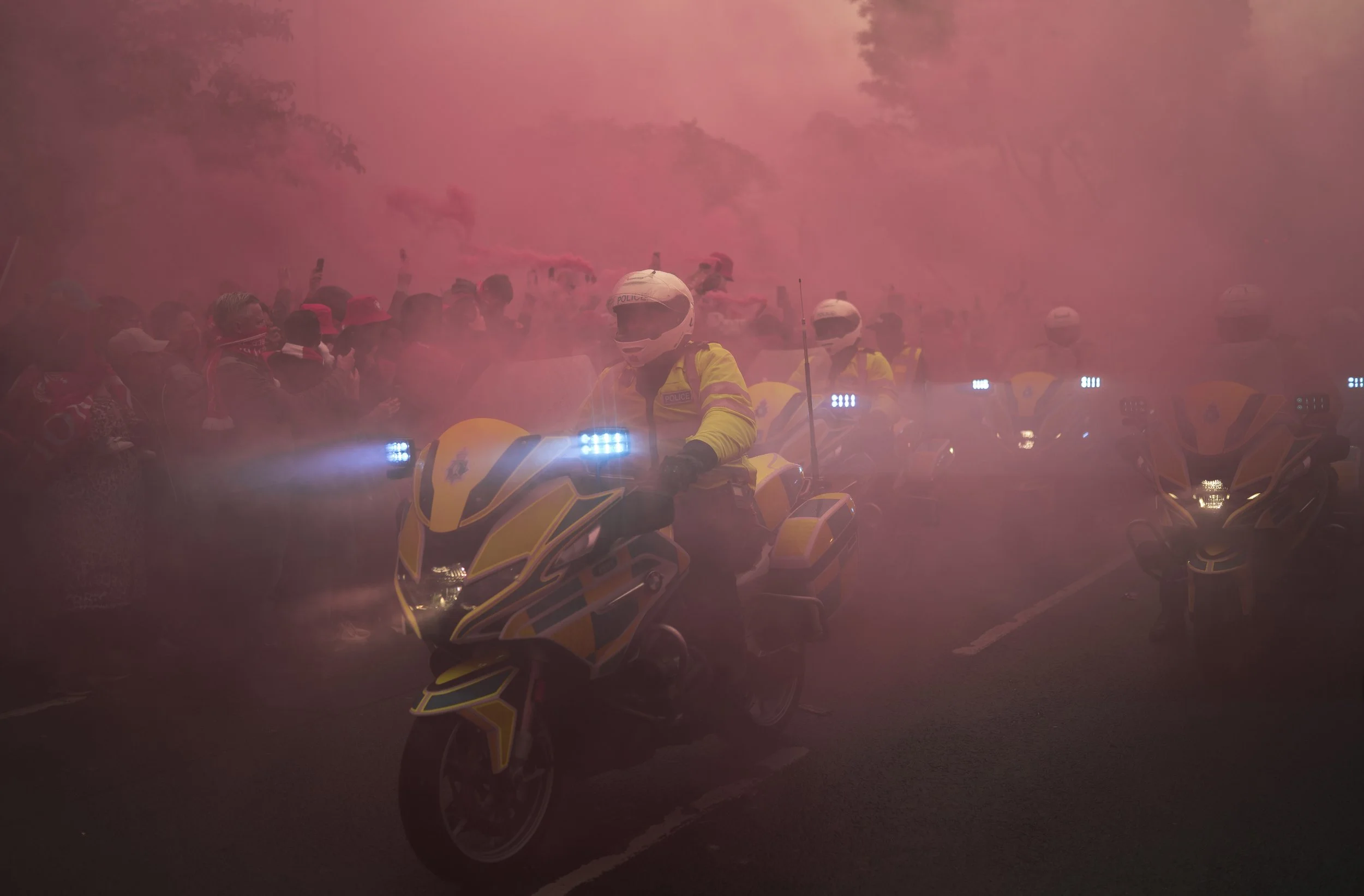 Police officers on yellow motorcycles surrounded by a crowd of people with pink smoke in the background.