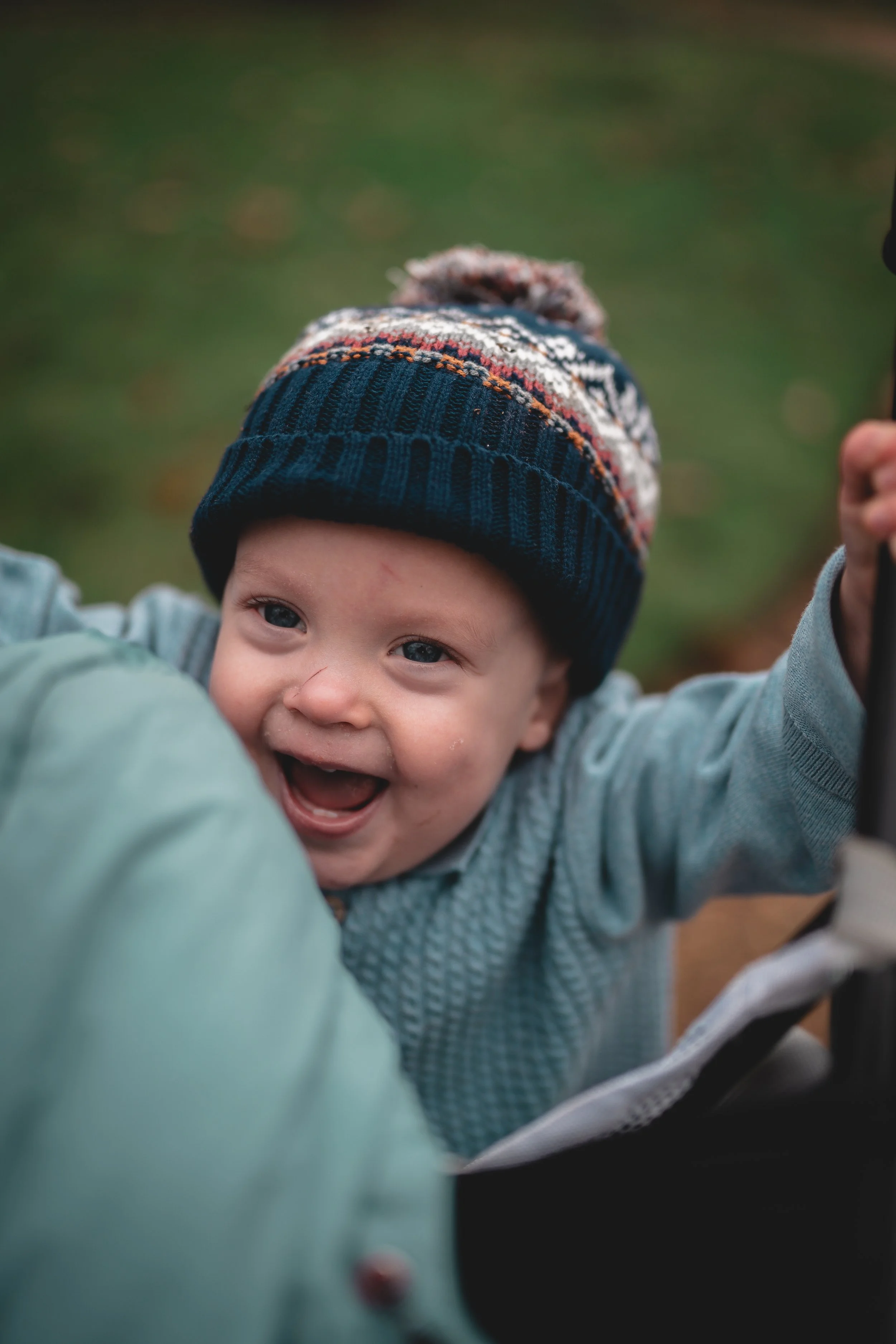 Close-up of a smiling baby wearing a festive knit hat, held by a person in a green jacket, outdoors with blurred greenery in the background.