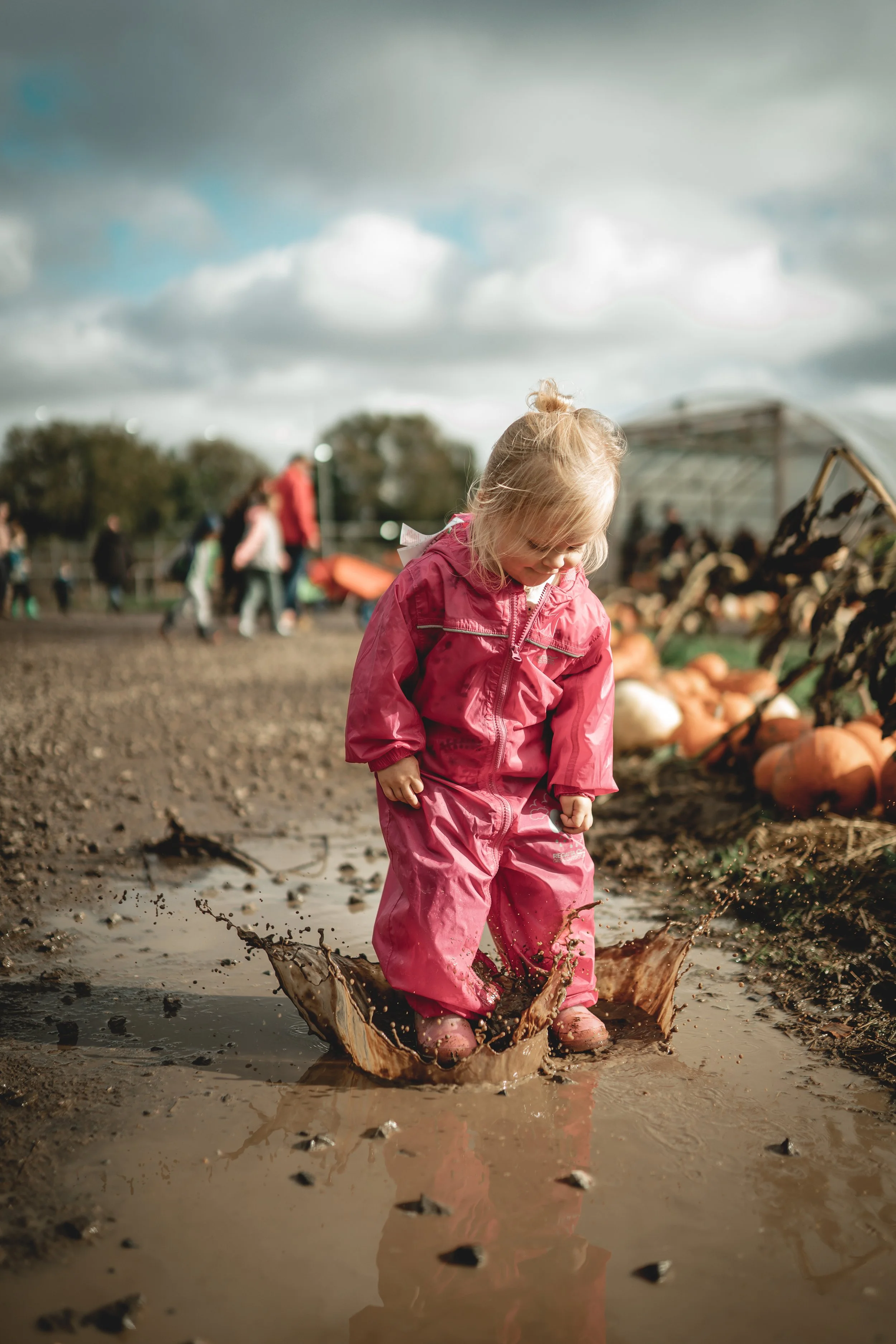 A young girl in a pink raincoat and boots jumping in a muddy puddle at a farm or pumpkin patch with other people and pumpkins in the background.