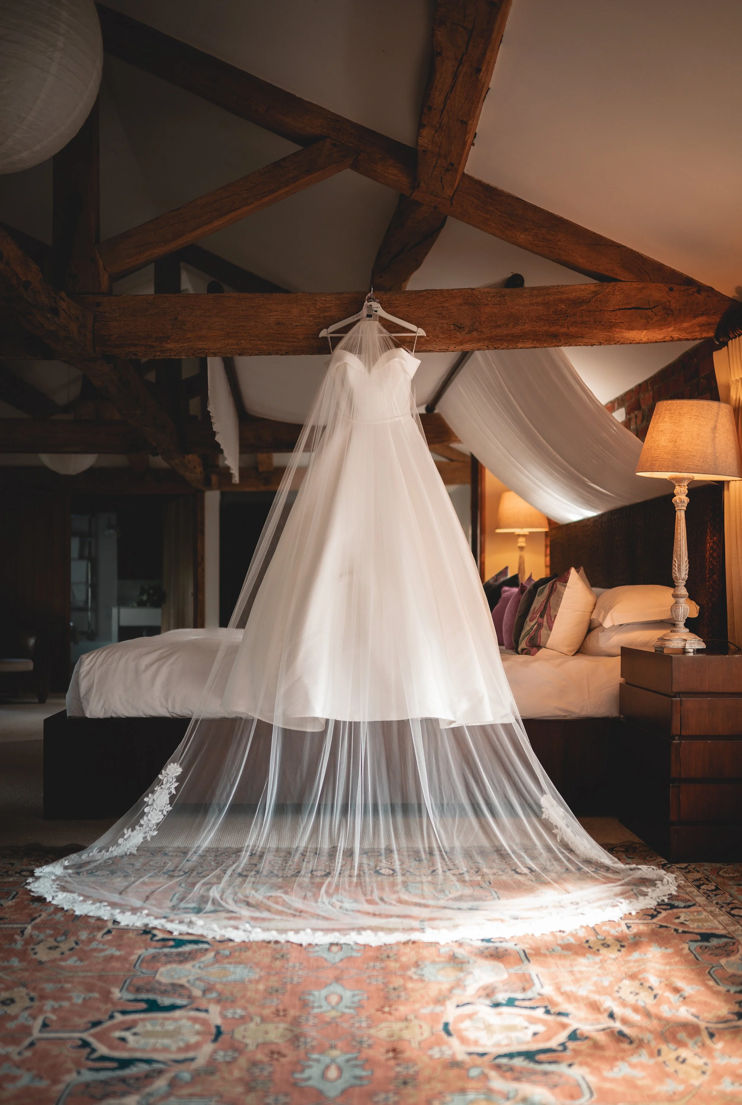 A white wedding dress hanging from a beam in a bedroom with wooden beams, lamps, and a quilted bed with pillows.