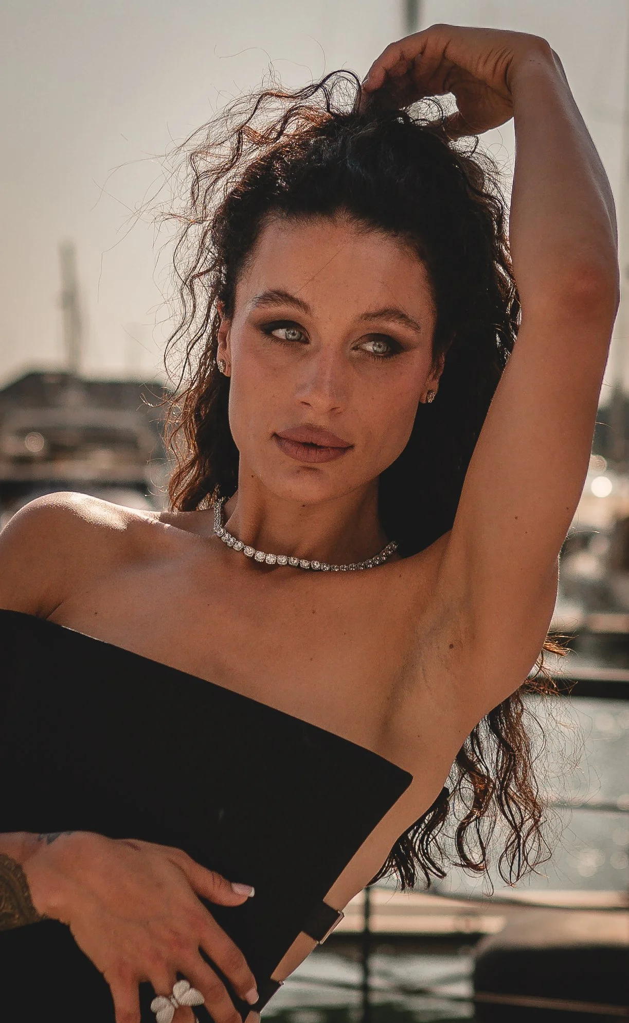A woman with long curly dark hair and light blue eyes, wearing a black strapless dress, a pearl necklace, and earrings, standing outdoors near a body of water, with her arm raised above her head.