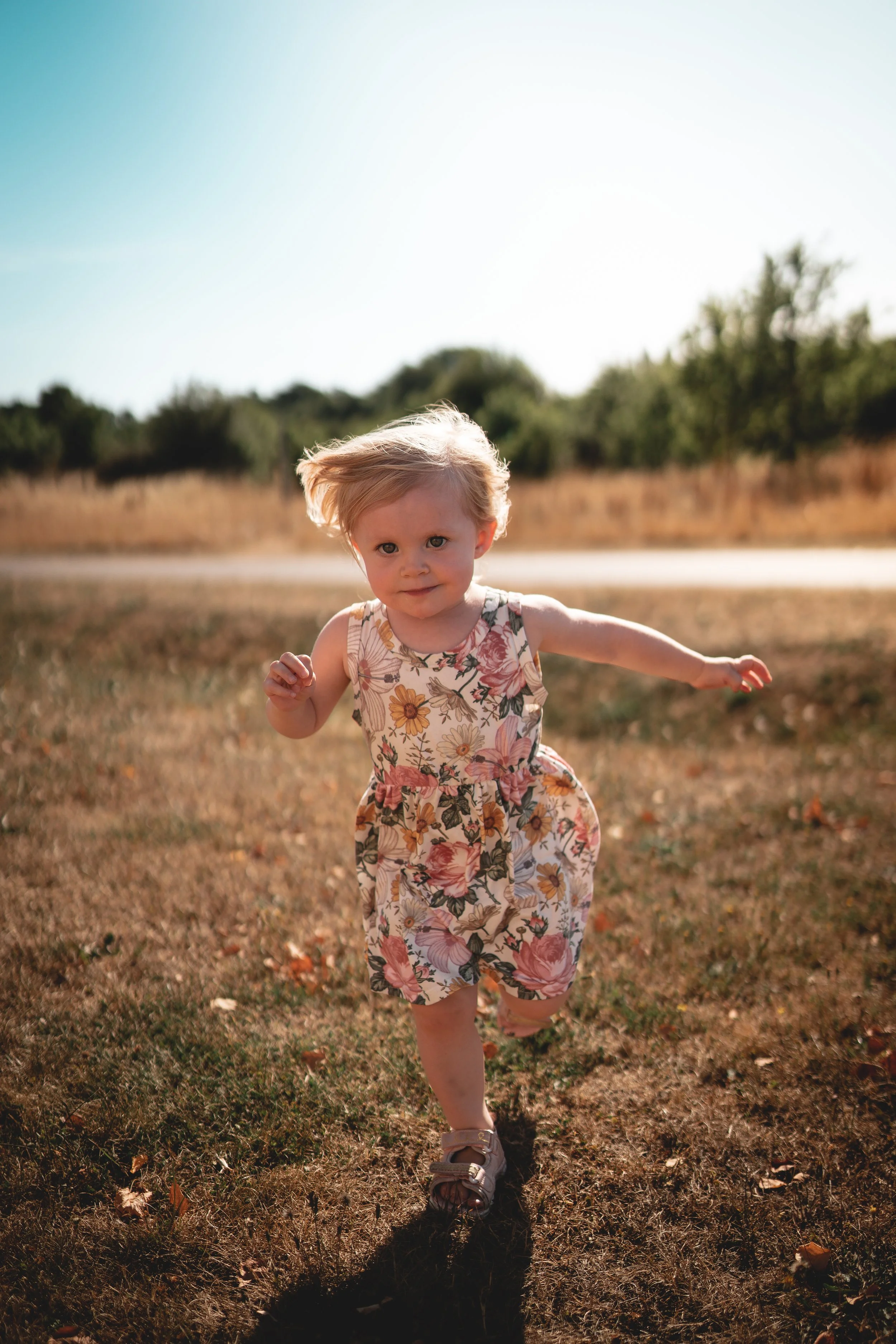 A young girl with blonde hair running through a grassy field on a sunny day, wearing a floral dress and sandals.