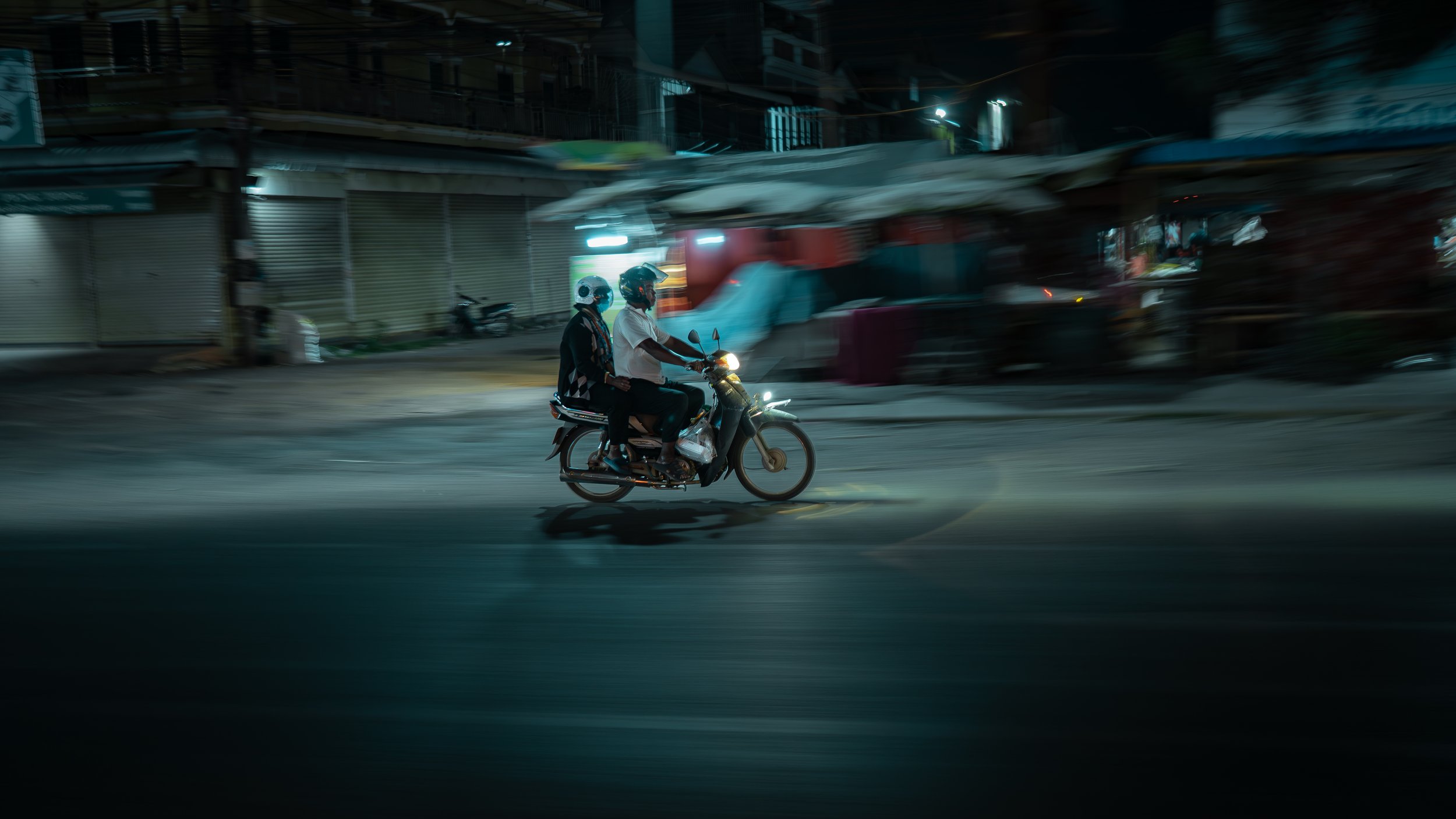 Two people riding a motorcycle at night on an urban street with closed shops and blurry background.