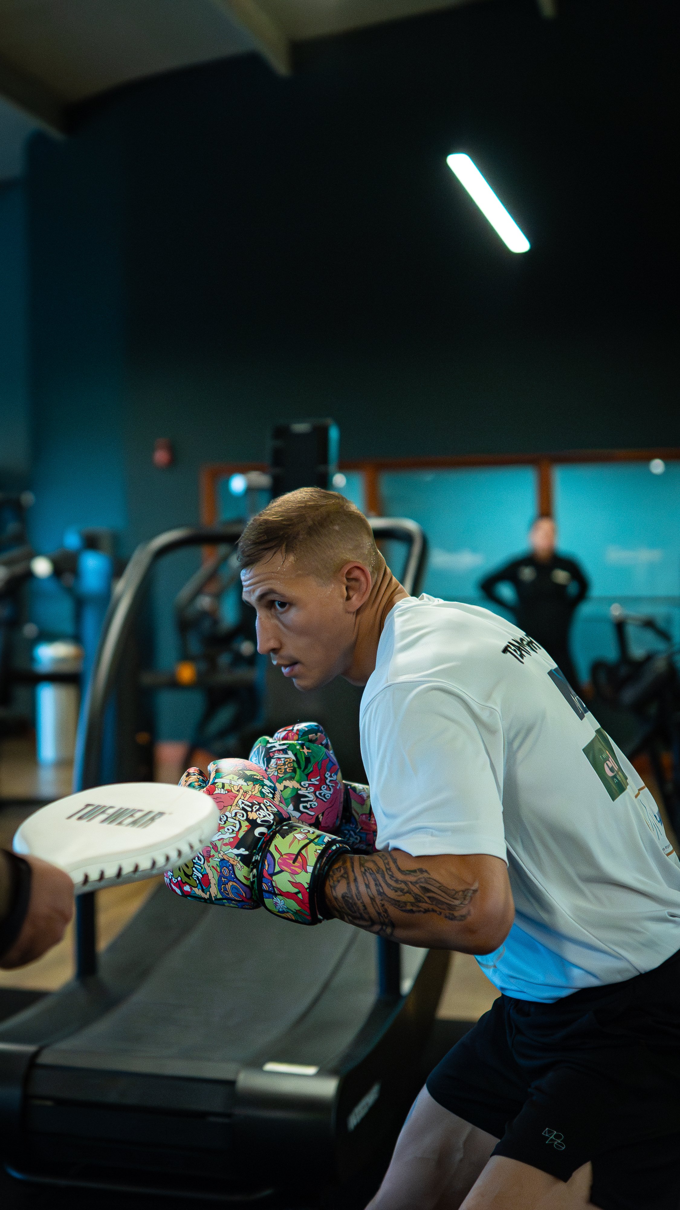 A young man in a gym wearing boxing gloves, leaning on a treadmill, with a trainer in the background.