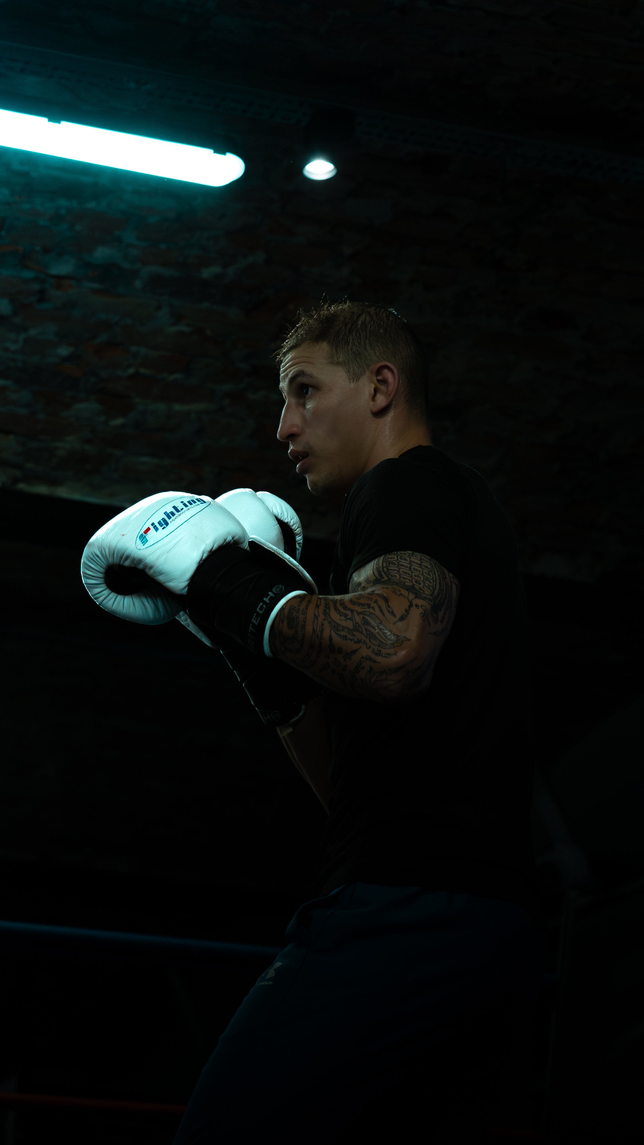 A young man with tattoos on his arm wearing boxing gloves, standing in a dimly lit boxing gym, preparing to spar or train.