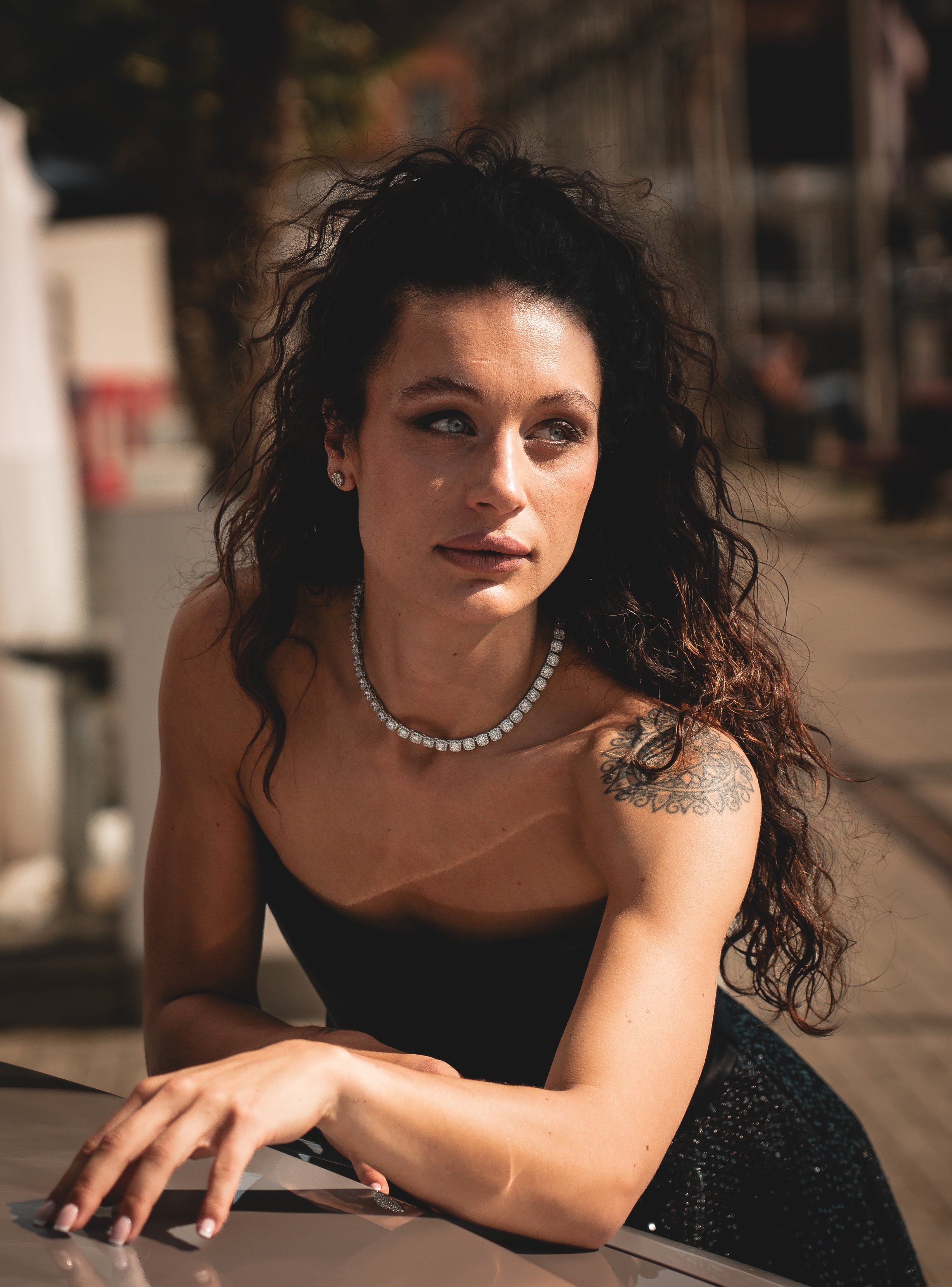 A woman with curly dark hair, wearing a black strapless top and diamond necklace, leaning on a car with an outdoor background.