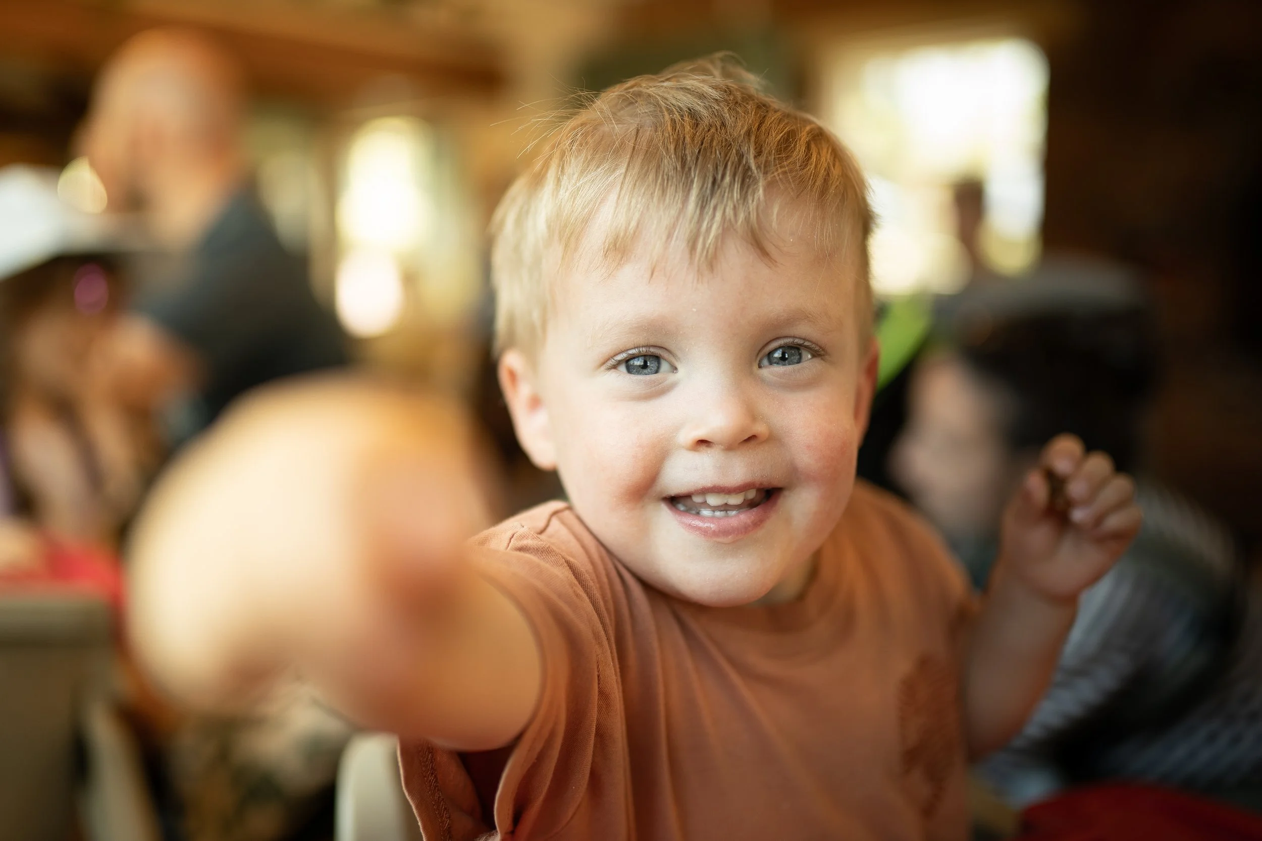 A young boy with blue eyes and a big smile pointing at the camera in a busy indoor setting.