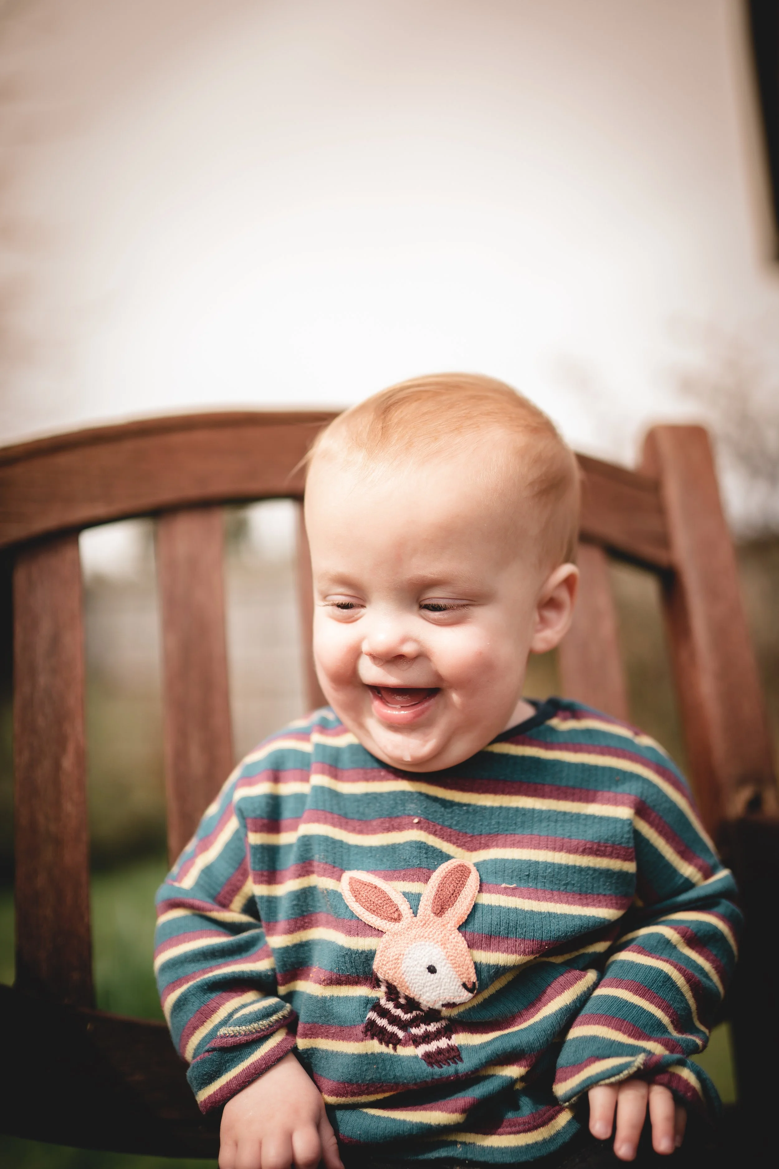 A young boy with blonde hair smiling and sitting on a wooden bench outdoors, wearing a striped sweater with a bunny patch.