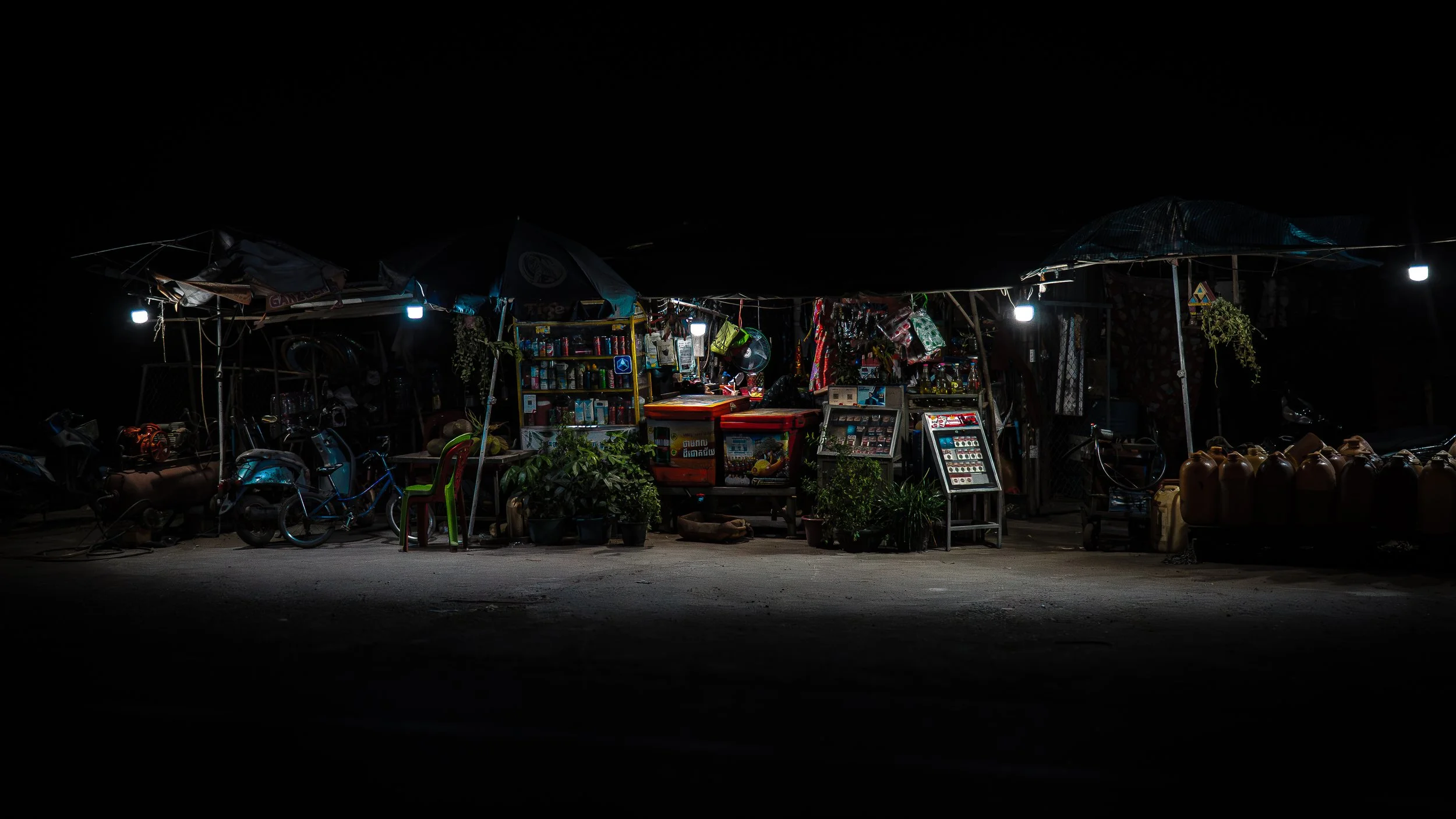 A small, outdoor convenience store at night, illuminated by a few bright lights, with shelves of snacks and beverages, some bicycles parked outside, and various potted plants around the shop.