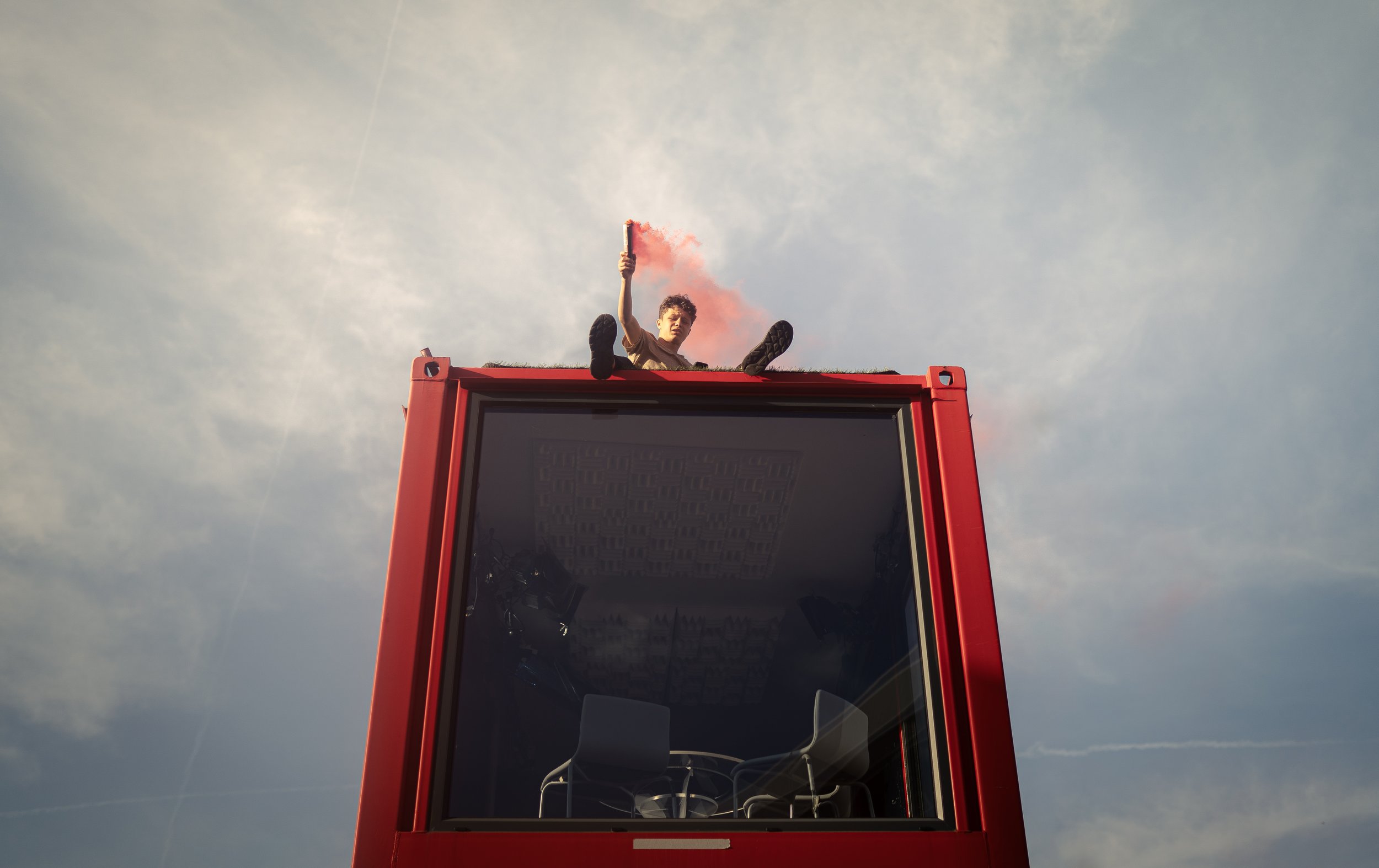 Person sitting on top of a red structure holding a smoke flare emitting pink smoke, with an interior view of an office or conference room visible through the glass below.