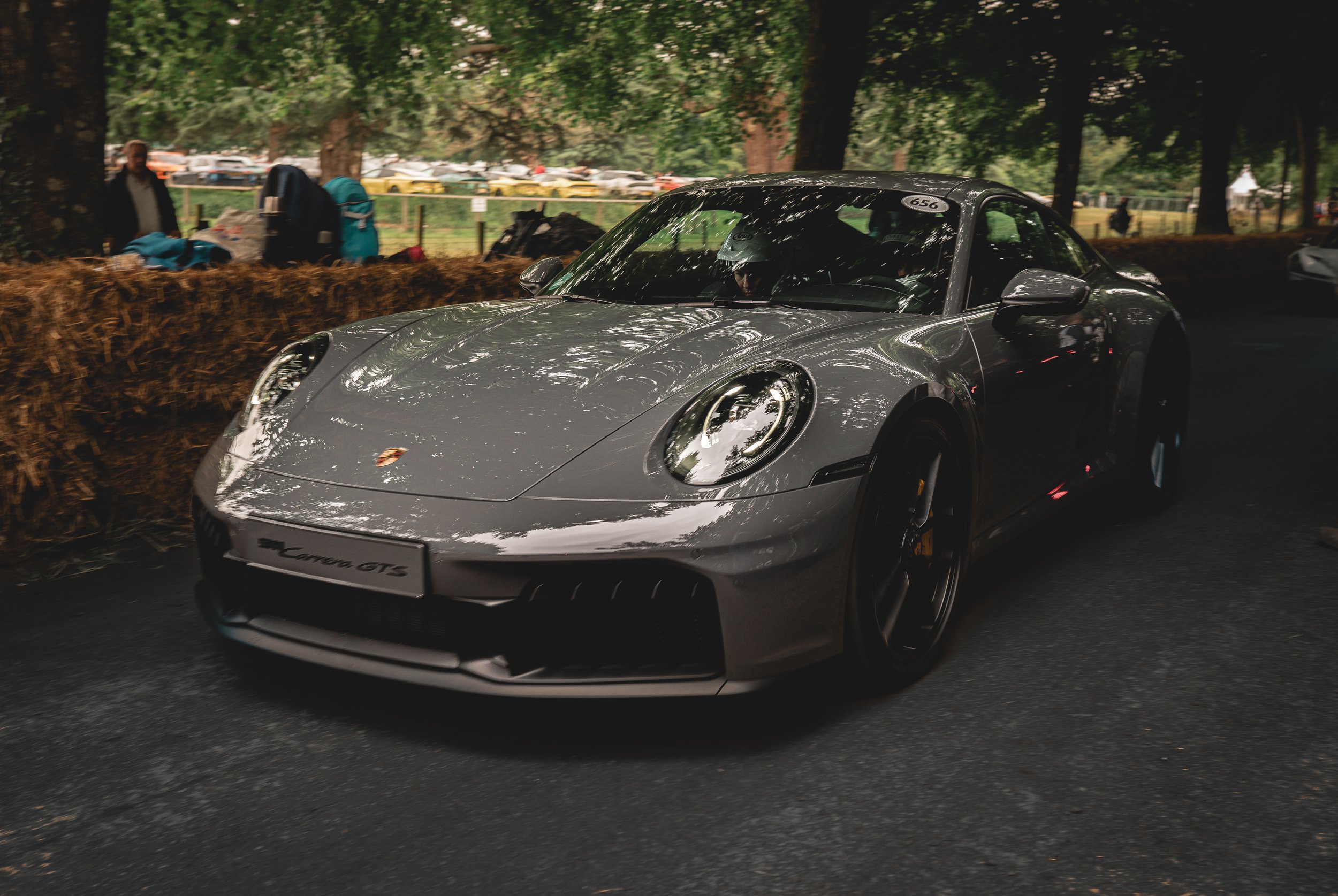 A silver Porsche 911 Carrera GTS sports car on a dark road, with trees and spectators in the background at a car show or event.