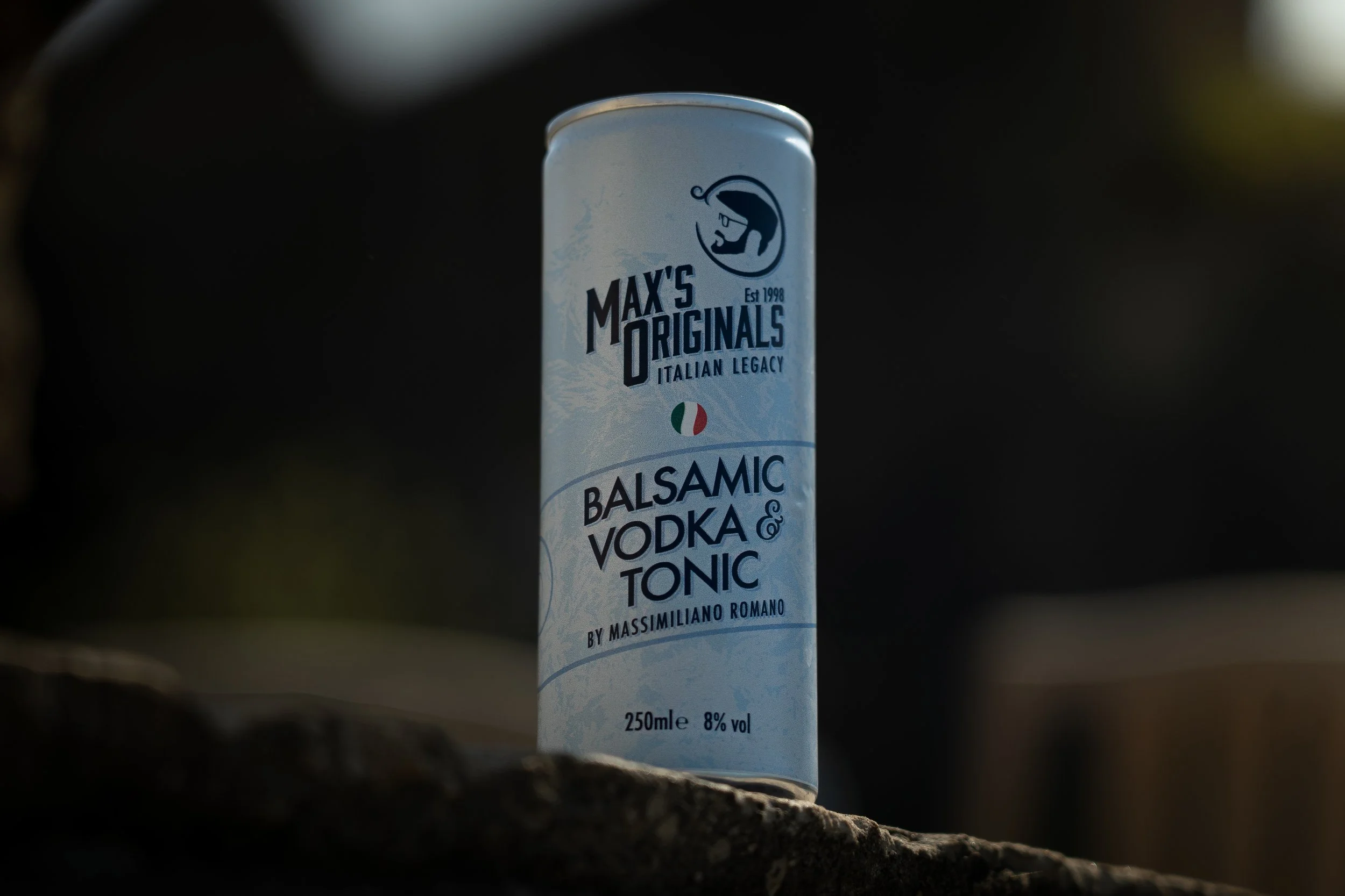 Can of Max's Originals Balsamic Vodka & Tonic beverage sitting on a surface outdoors, with a dark blurry background.