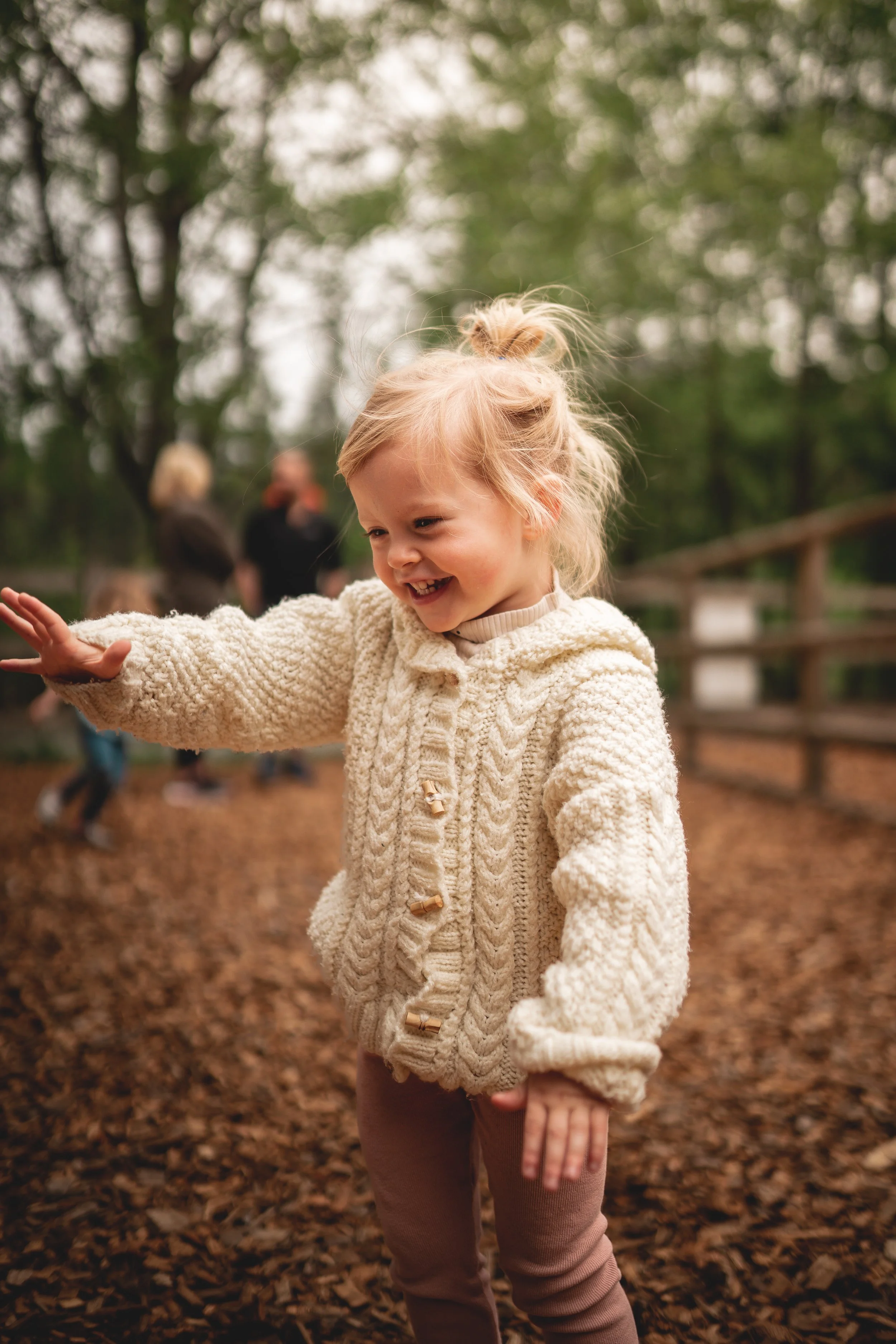 A young girl with blonde hair in a ponytail, wearing a cream-colored knitted sweater, smiling and reaching out in an outdoor wooded area.