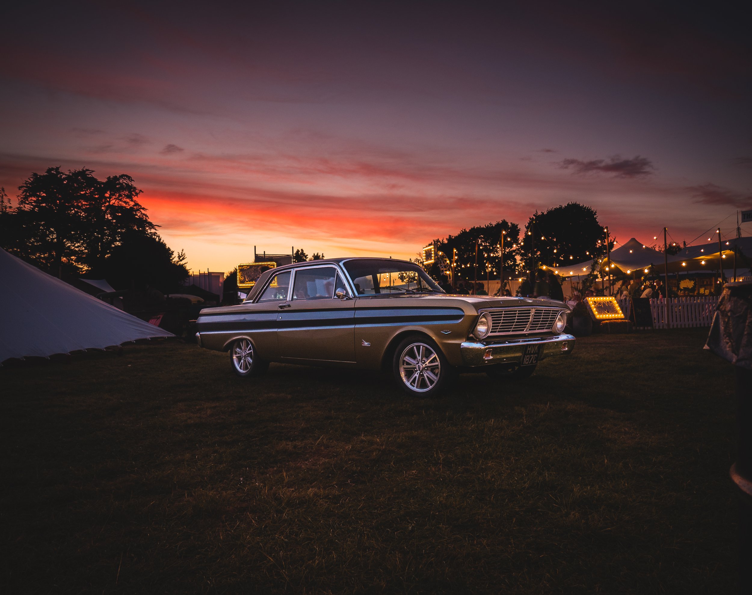 A vintage car parked on a grassy field at sunset with carnival tents and string lights in the background.