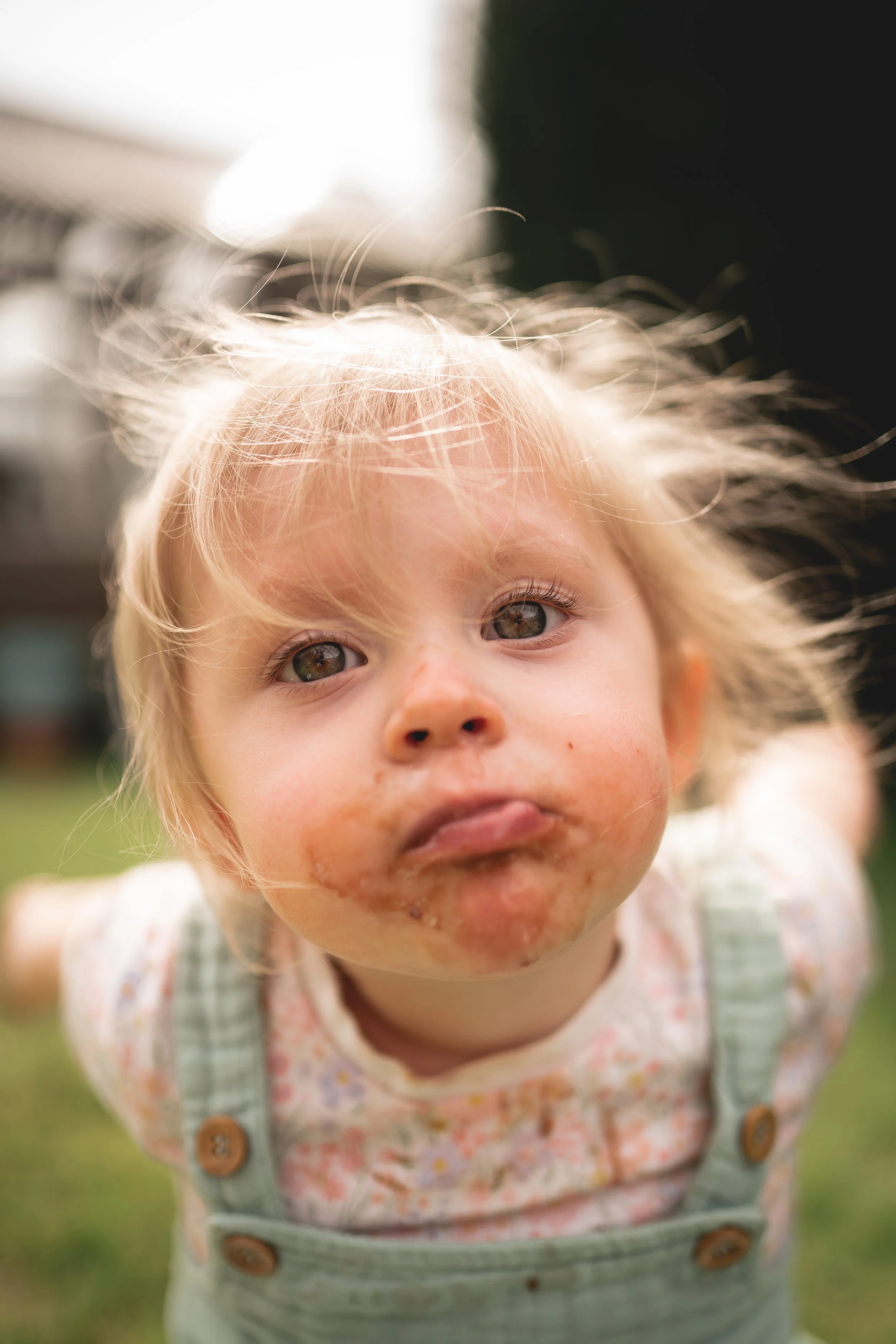 Close-up of a young girl with messy blonde hair, dirt smudges on her face and around her mouth, looking into the camera with a curious expression, outdoors on a grassy area.