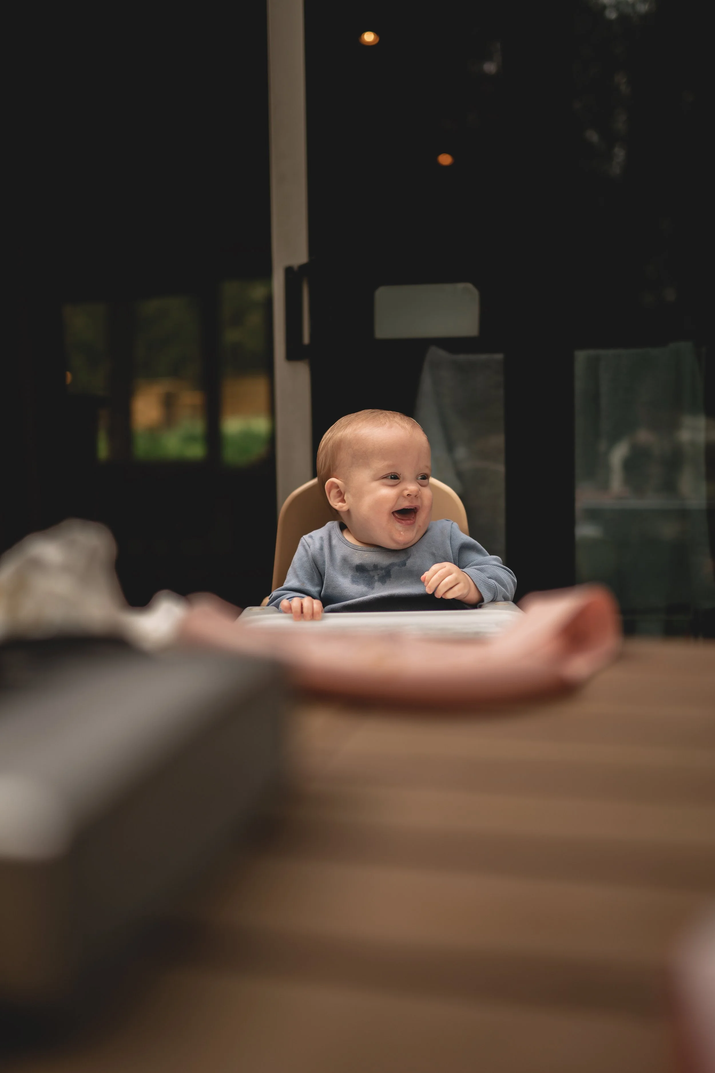 A baby with short hair and a gray shirt sitting in a high chair, smiling and laughing, at a table indoors, with a blurred background.
