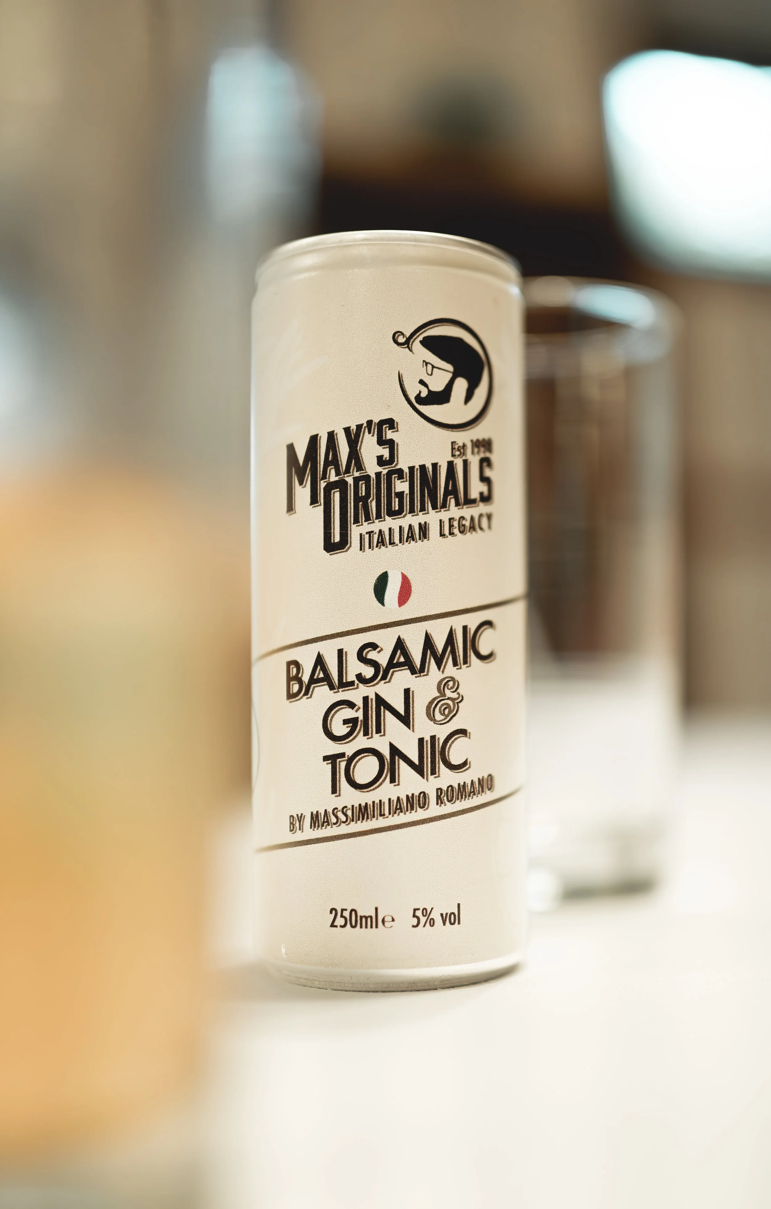 A can of Max's Originals balsamic gin and tonic, featuring a black and white label with an illustration of a hand holding a glass and Italian flag colors, placed on a white surface.