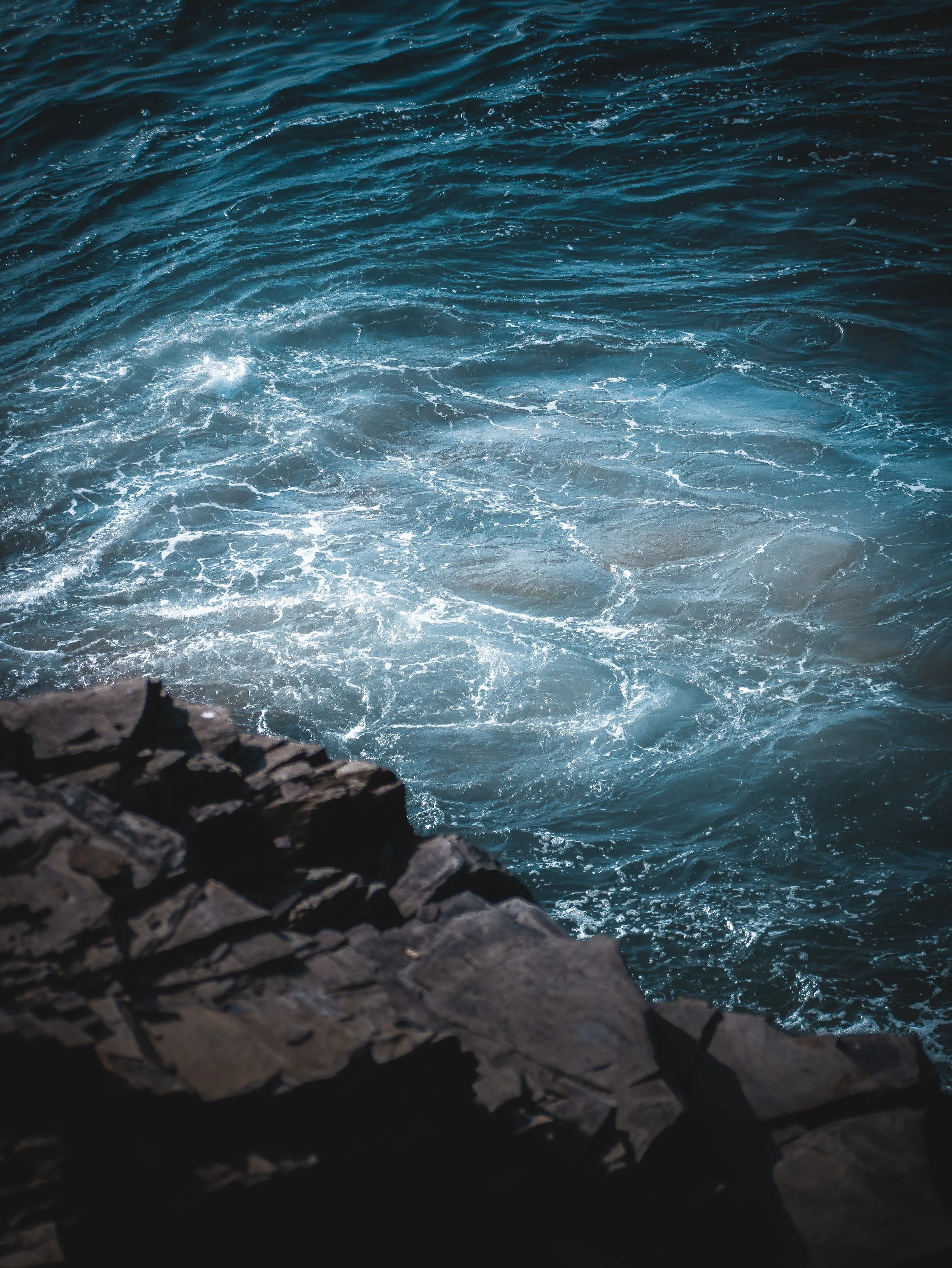 Ocean waves seen from the rocky shoreline, with white foam and churning water.