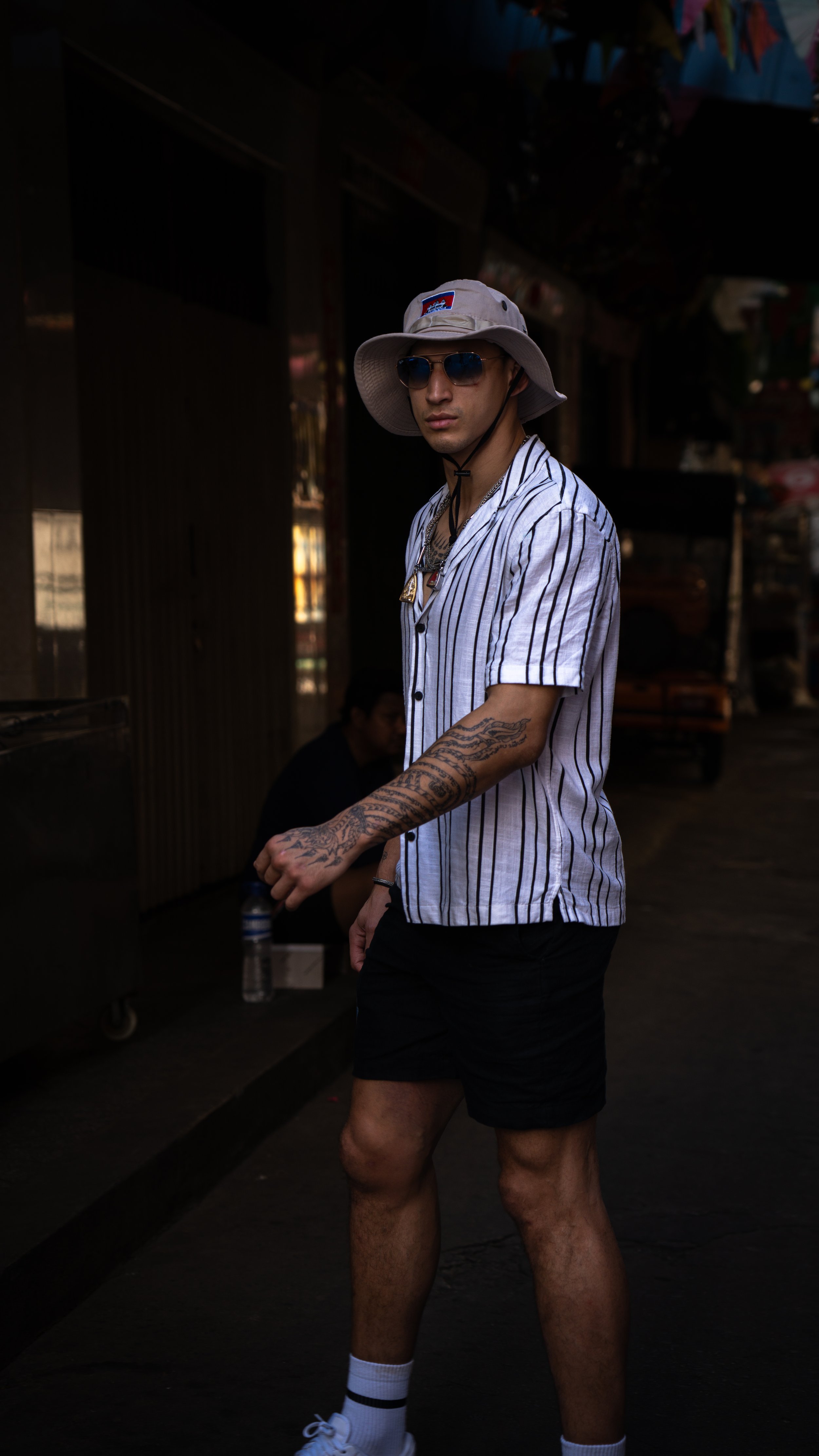 A young man standing outdoors at night, wearing sunglasses, a wide-brimmed hat, a striped short-sleeve shirt, black shorts, white socks, and sneakers. He has tattoos on his left arm and some jewelry around his neck.