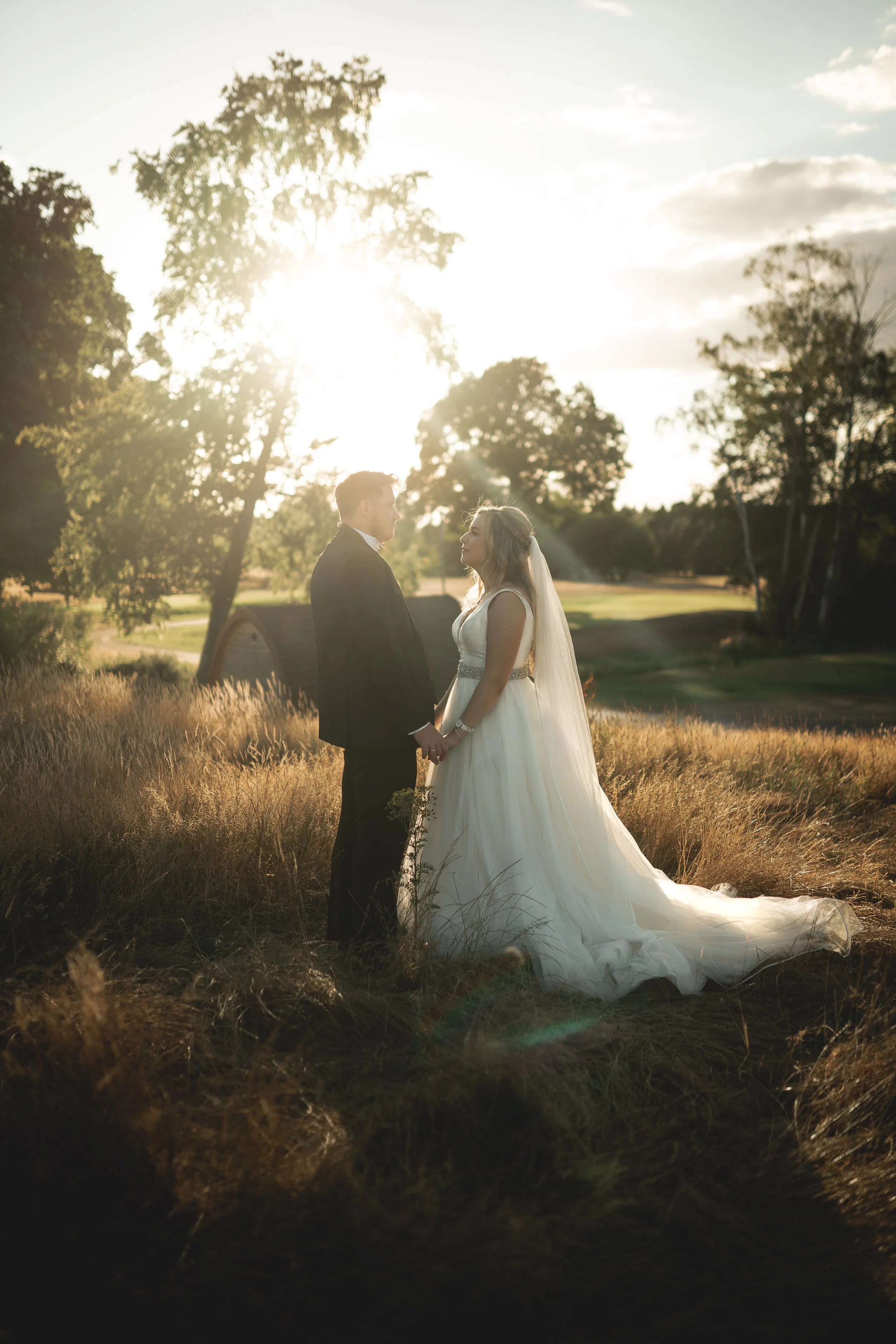 A bride and groom holding hands and looking at each other outdoors during sunset, surrounded by tall grass and trees.