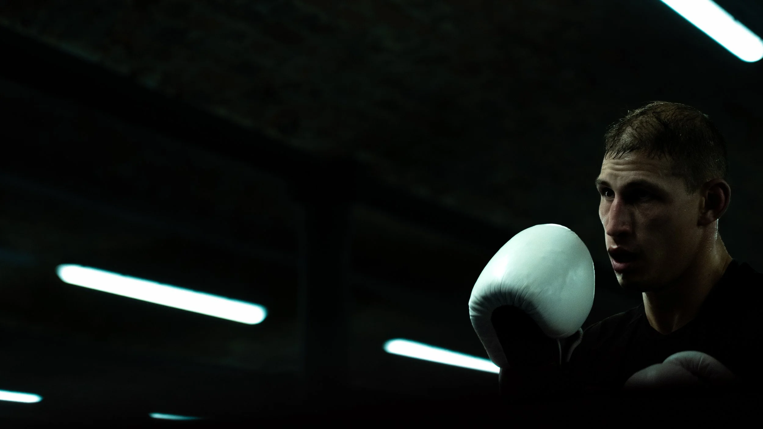 A male boxer in a dark gym, wearing black gloves, preparing for a fight with focused expression.