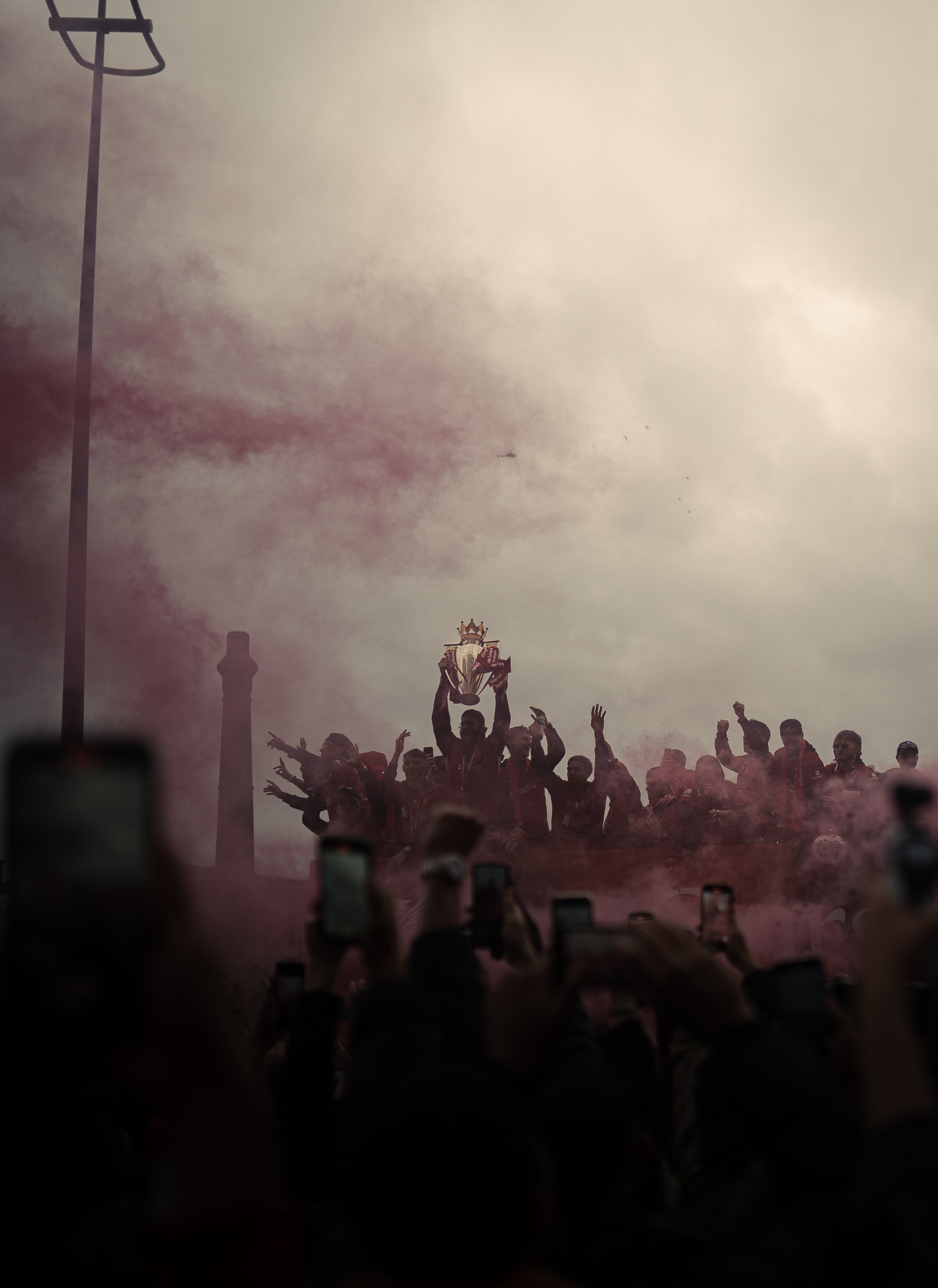 A sports team celebrates on a podium, holding a trophy, surrounded by smoke or fog, with multiple fans taking pictures and cheering in the foreground. The background features a cloudy sky with a tall lamppost and a smokestack.