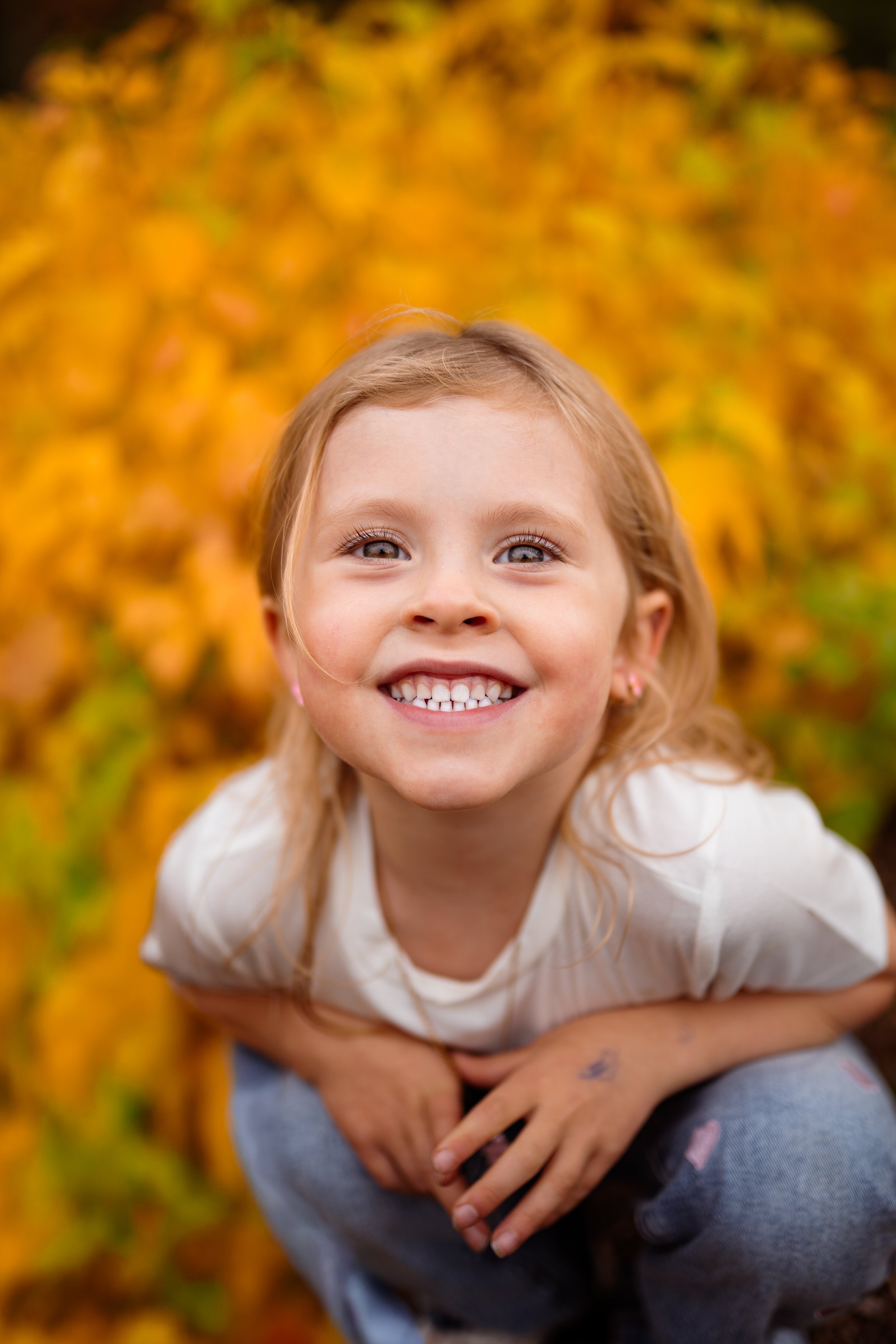 Close-up of a smiling young girl with blonde hair and blue eyes, sitting outdoors in front of autumn foliage with yellow and orange leaves.