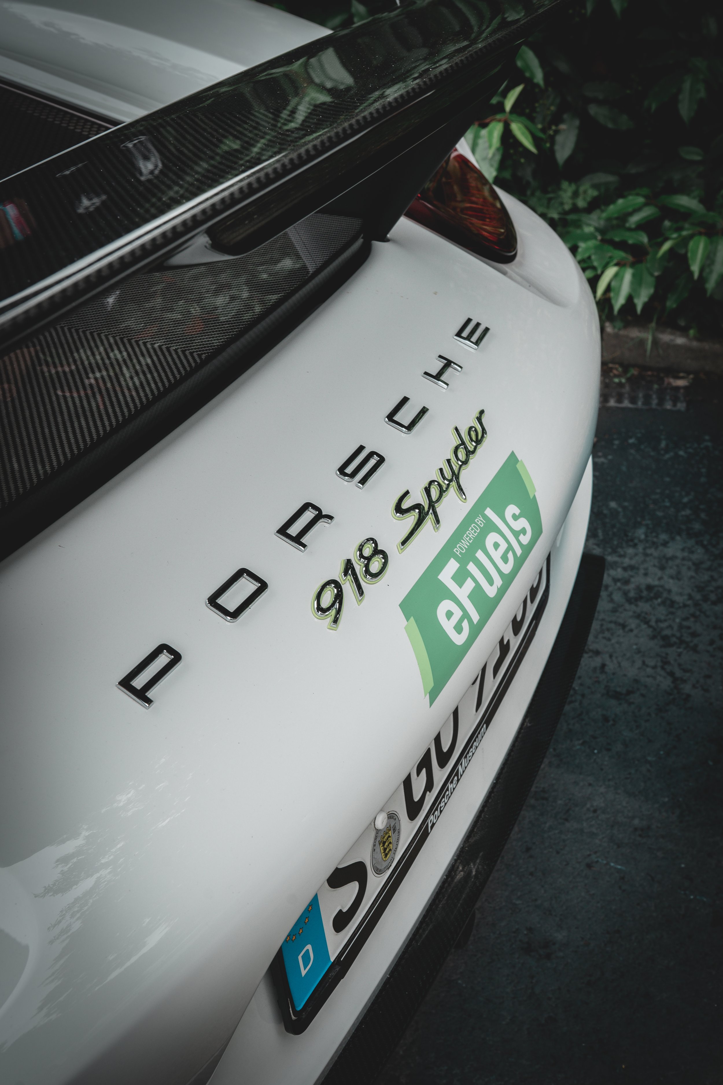 Close-up of the rear of a white Porsche 918 Spyder sports car with sponsor logos, including e Fuels, and a black carbon fiber rear wing.
